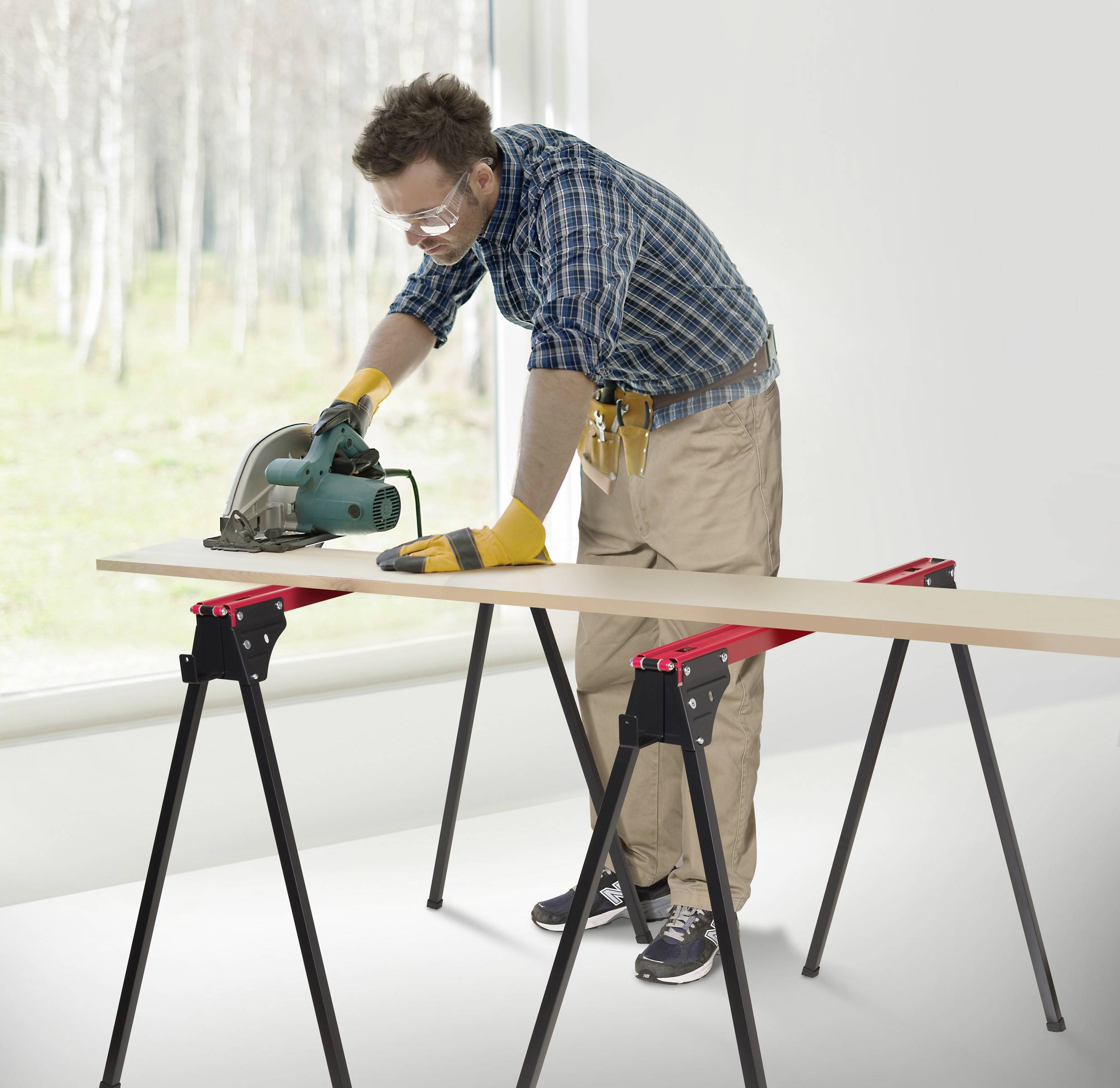 A man wearing safety goggles and gloves uses a circular saw to cut a wooden plank on sawhorses in a bright room with a large window.