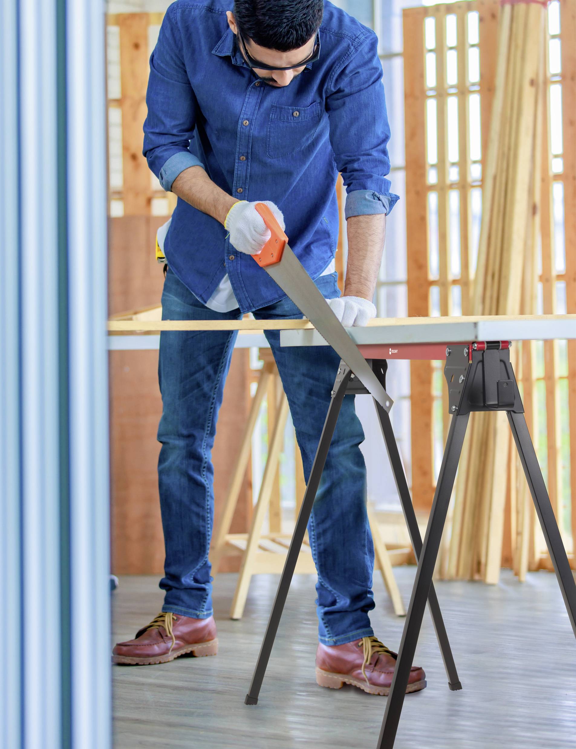 A person in a denim shirt and jeans is using a handsaw to cut a piece of wood supported by sawhorses, in a workshop setting.