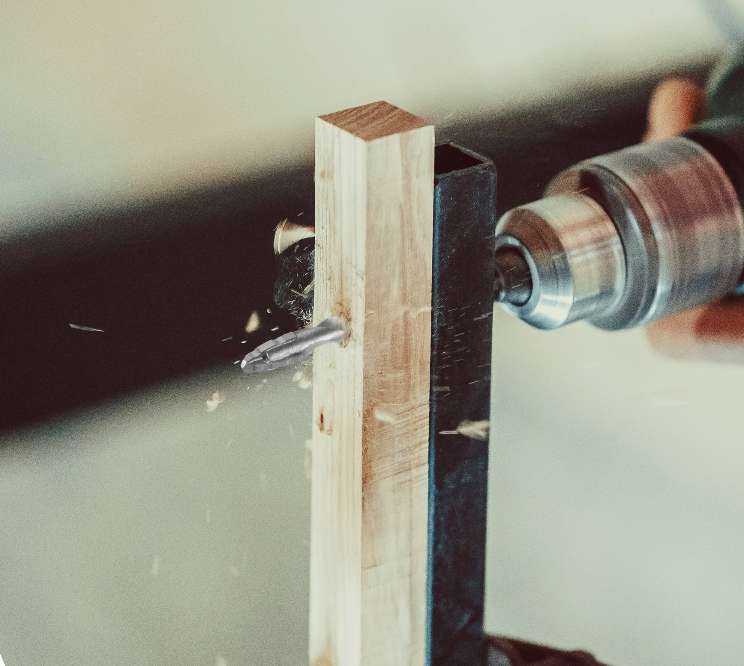 A power drill is boring a hole through a piece of wood, with wood shavings flying. The focus is on the rotating drill bit.