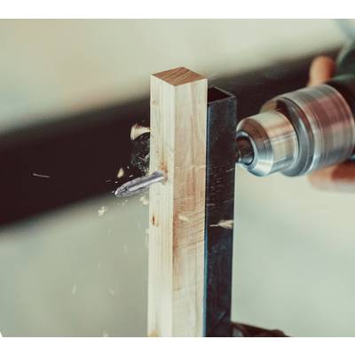 A power drill is boring a hole through a piece of wood, with wood shavings flying. The focus is on the rotating drill bit.