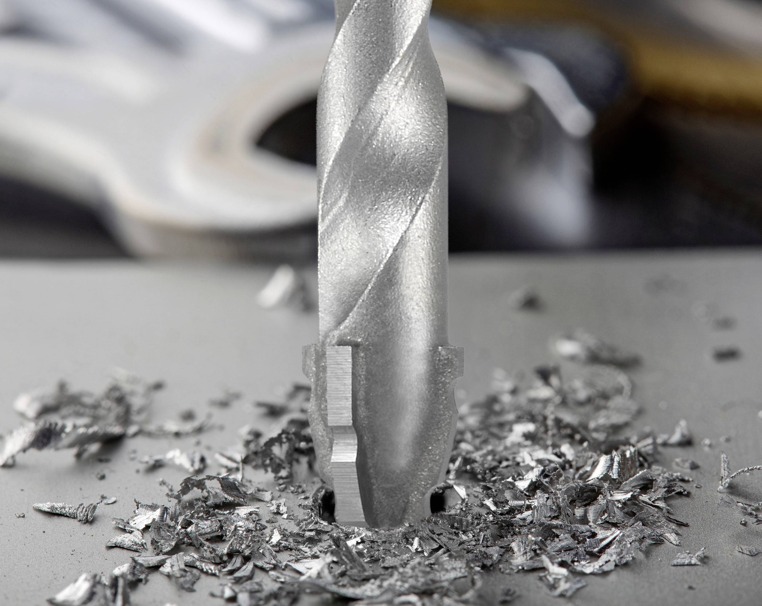 A close-up of a metal drill bit actively drilling into a grey surface, surrounded by scattered metal shavings.