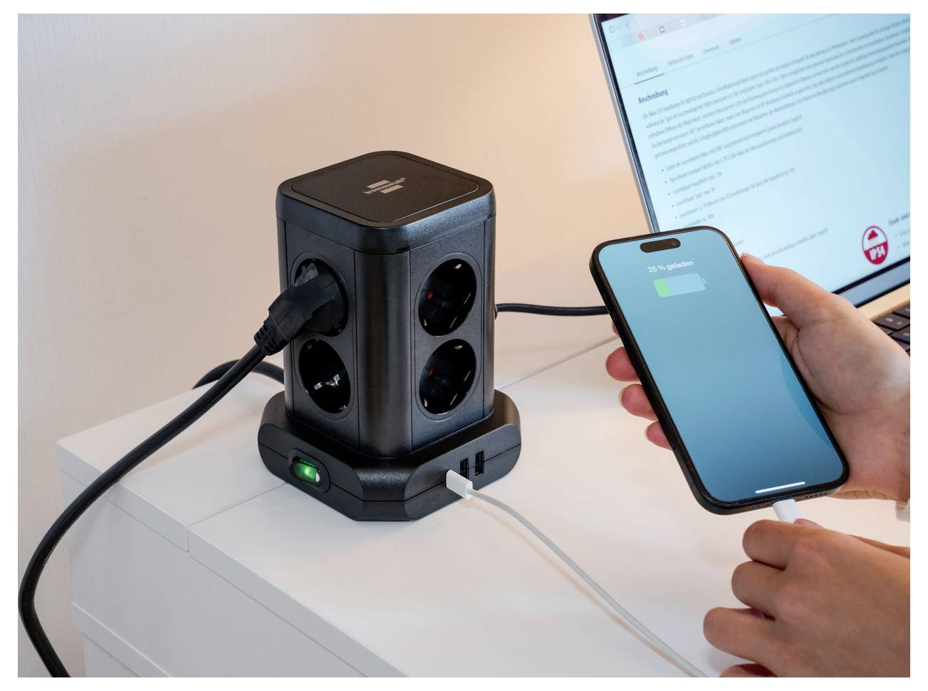 Hand plugging a smartphone into a cube-shaped desktop power station with multiple AC outlets and charging ports beside a laptop.