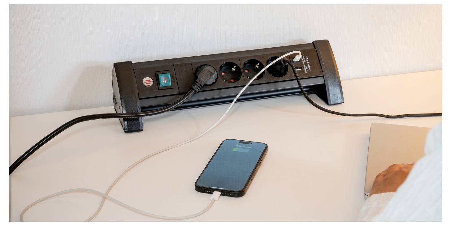 Smartphone charging on a desk via a black power strip with multiple plugs and cords.