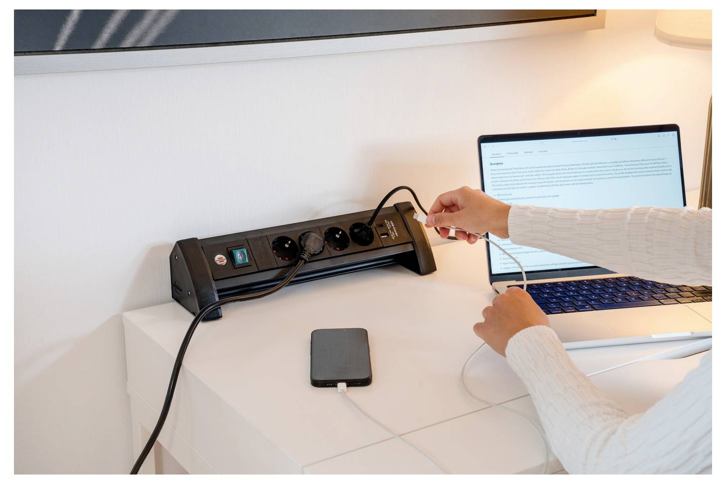 Person plugging a cable into a desk power strip beside a laptop and external drive on a white table.