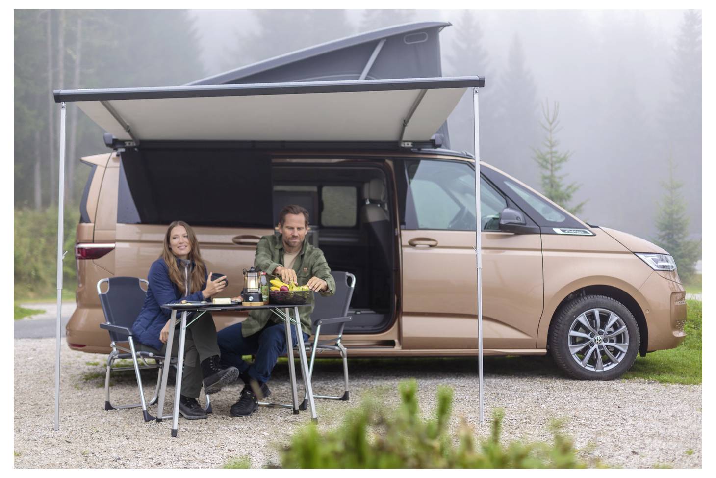 Couple sits at a folding table under a pop-up awning beside a tan camper van in a misty outdoor campsite.<br>