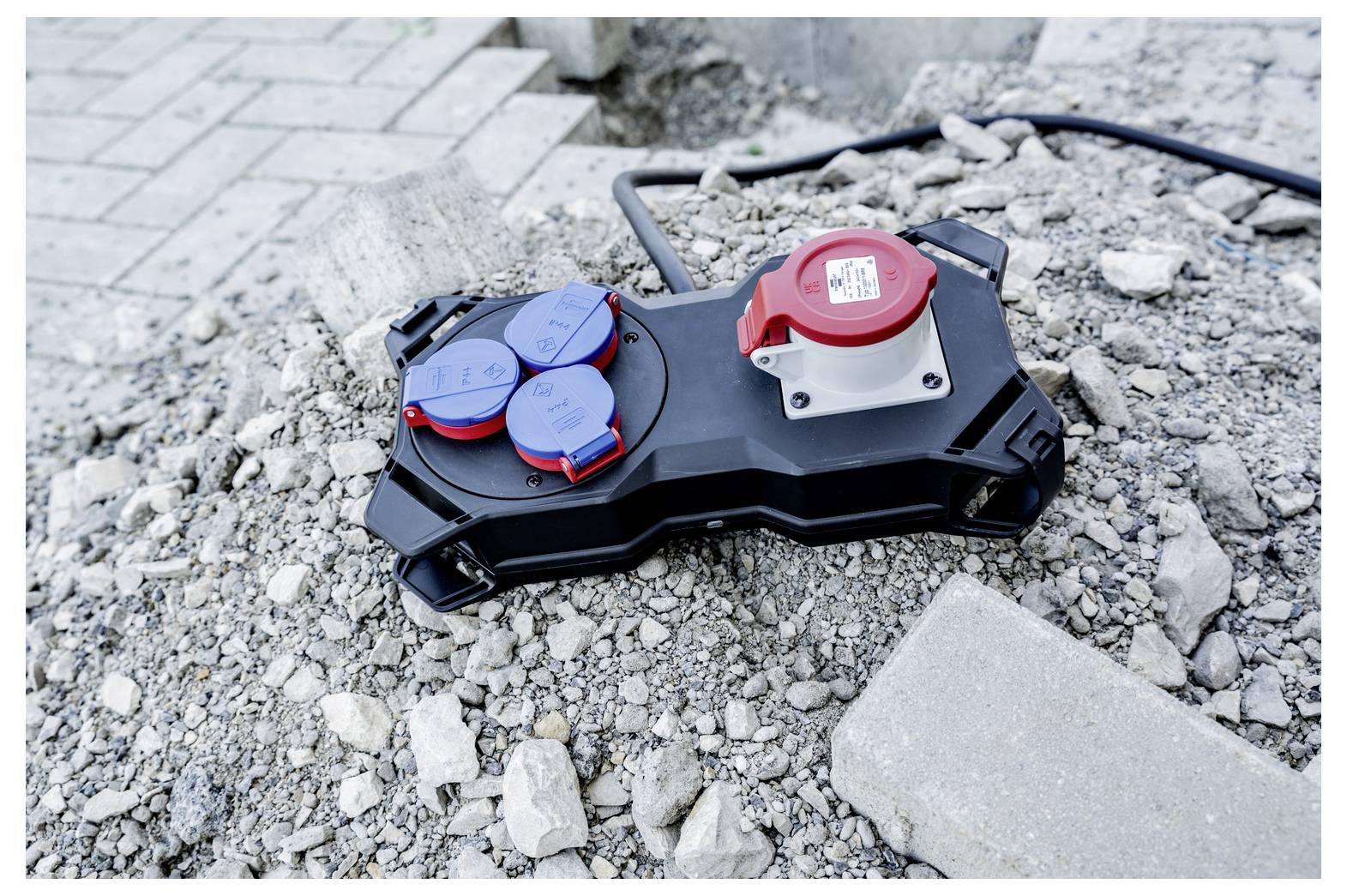 Portable power distribution box with red and blue outlets lying on gravel and construction debris near a paved surface.