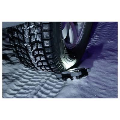 Close-up of a car tire with deep winter tread gripping snowy road, with a tire chain/tensioner visible near the wheel.