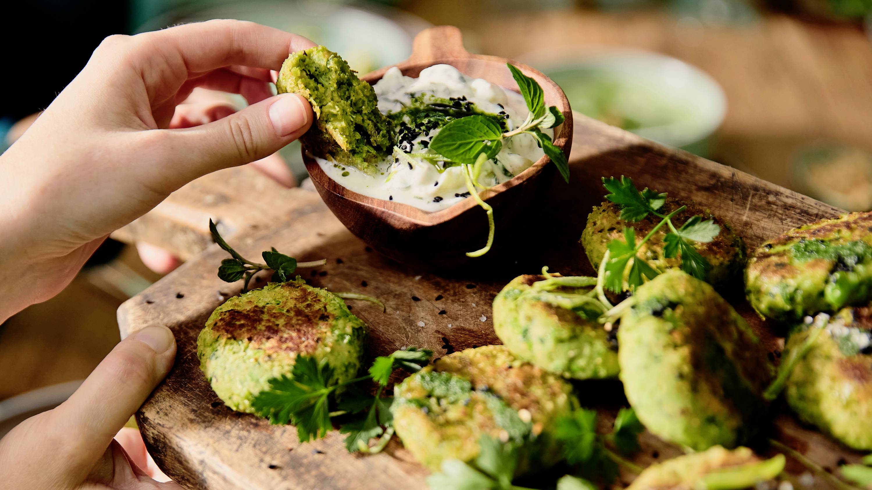 A person dips a green vegetable patty into a bowl of creamy white sauce on a wooden board with more patties and fresh herbs.