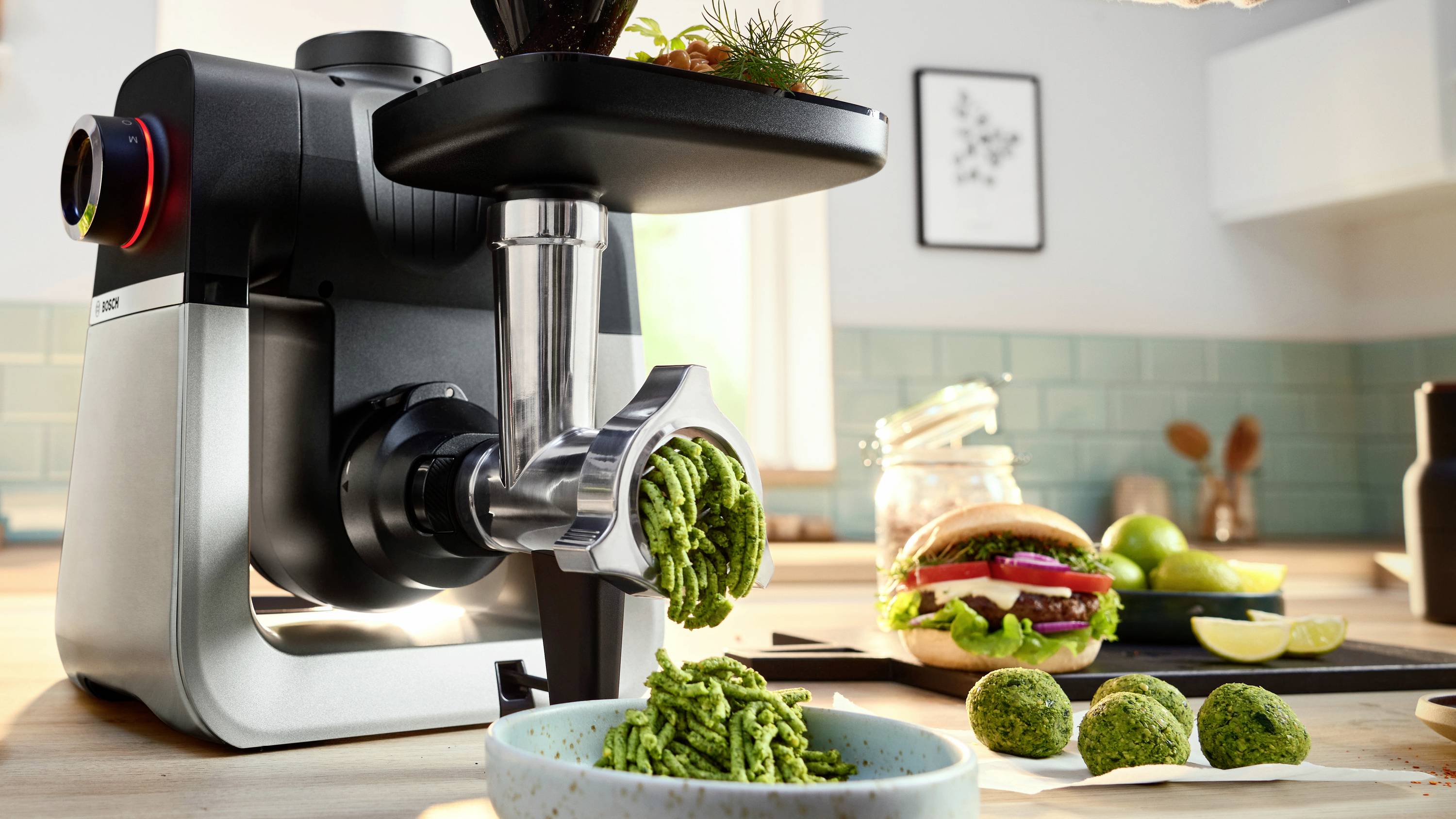 A kitchen scene with a meat grinder extruding green paste into a bowl. Ingredients and a sandwich are on the counter in the background.