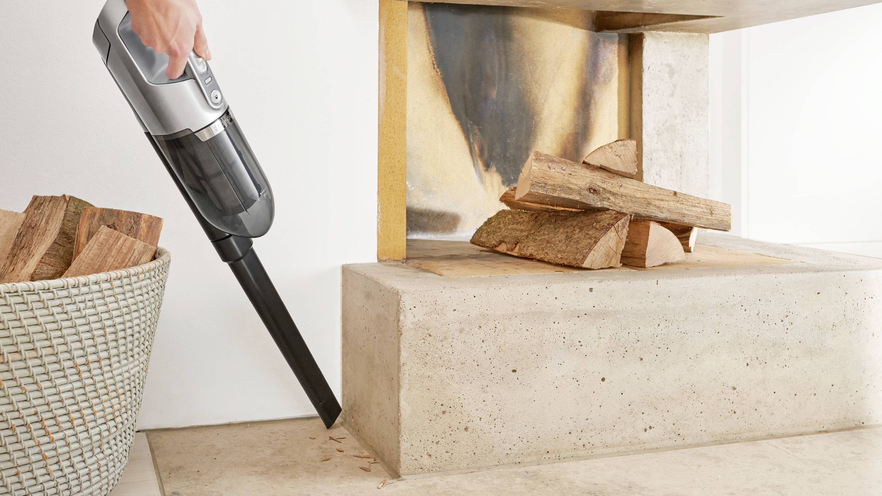 A person uses a cordless vacuum to clean dust around a fireplace with stacked firewood.