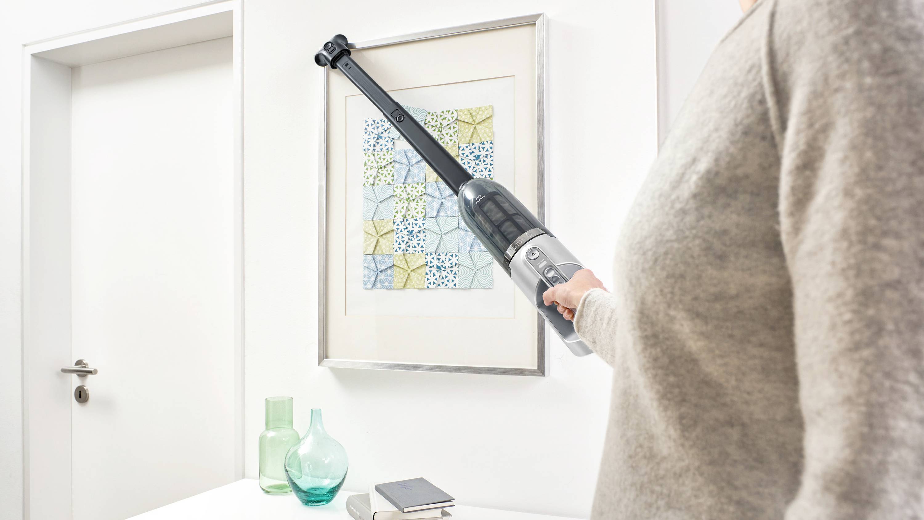 A person uses a long-handled vacuum cleaner to clean a picture frame on a white wall, with glass vases and a book nearby.
