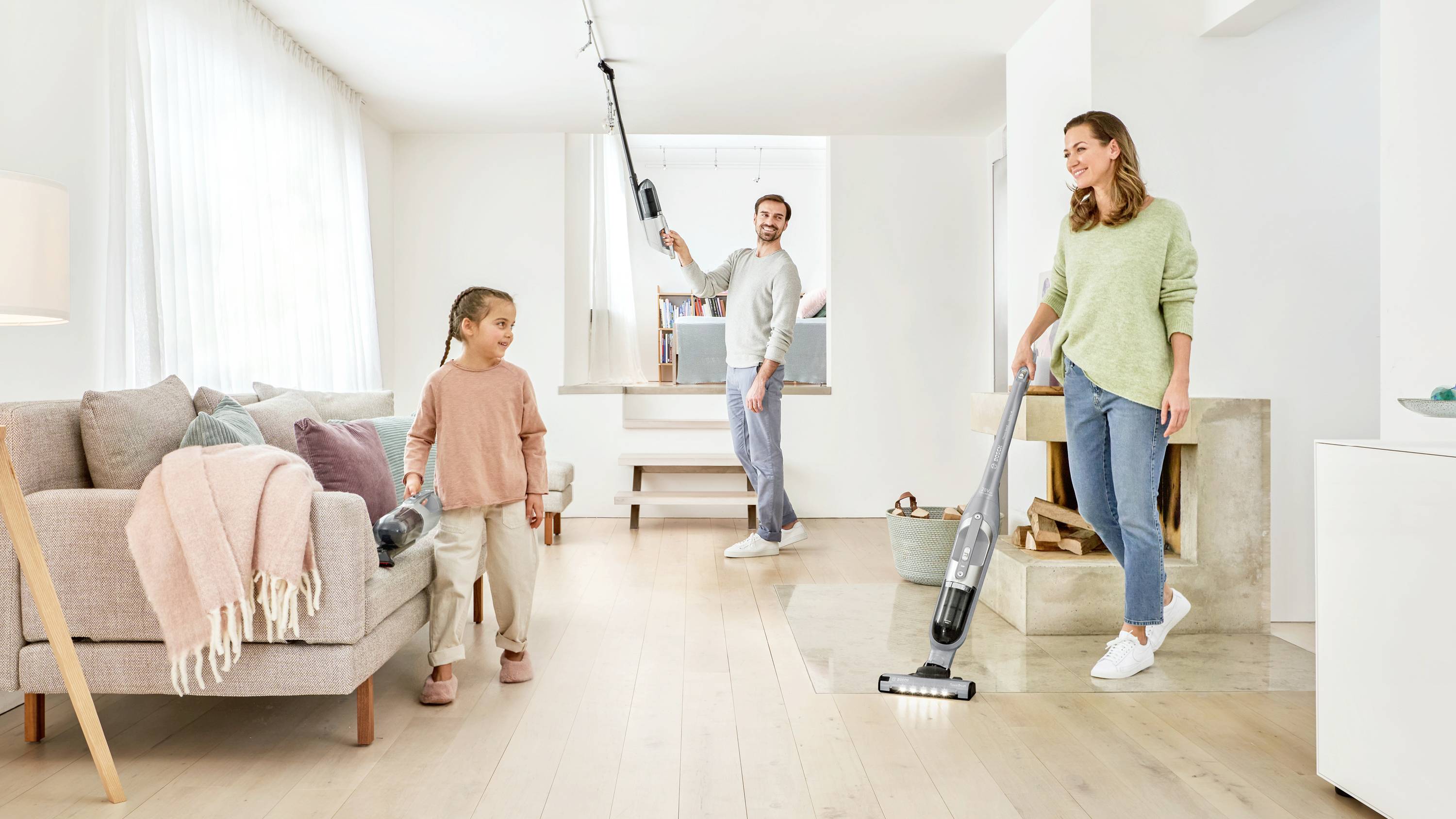 A family is using vacuum cleaners to clean a bright living room. A woman vacuums the floor, a man cleans the ceiling, and a child helps.