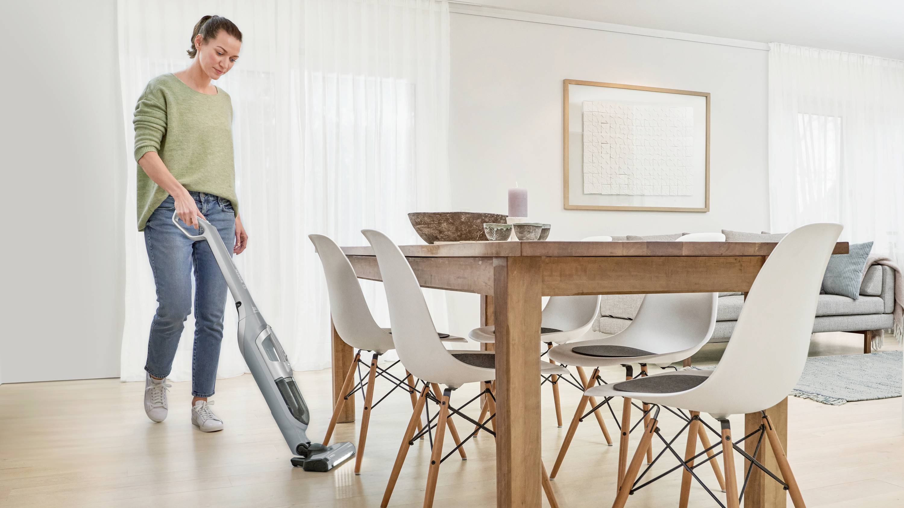 A person vacuuming a clean, modern dining area with wooden table and white chairs, conveying a sense of cleanliness and tidiness.