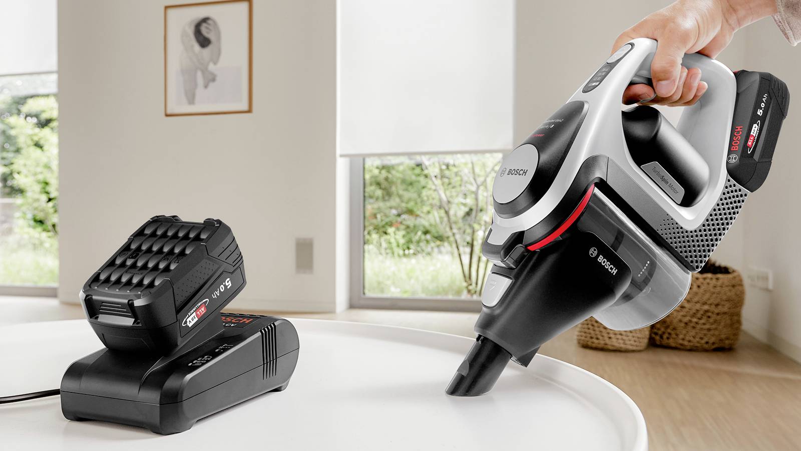 A hand holds a cordless vacuum cleaner above a table, near a charging dock. A window with a garden view is in the background.