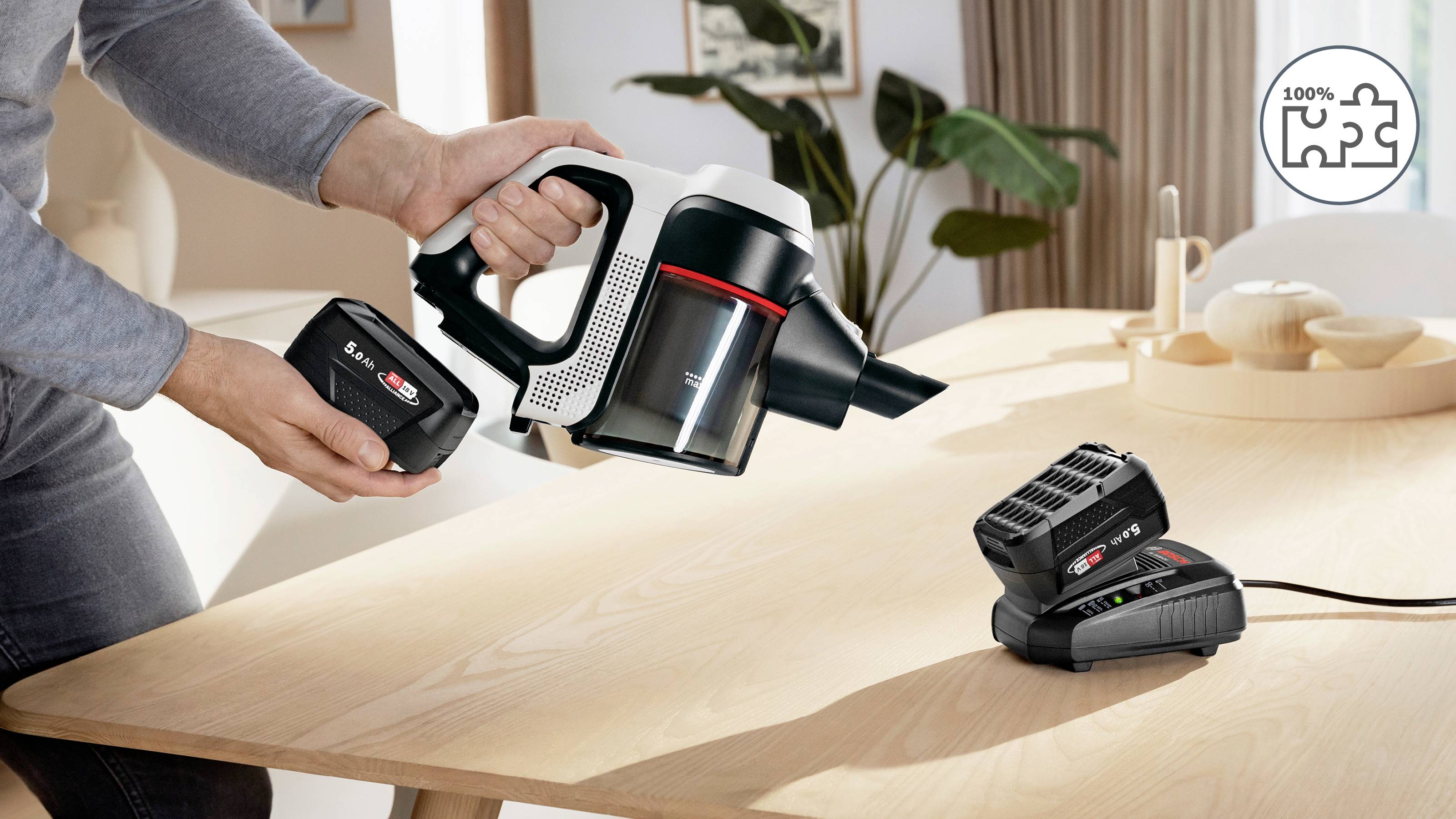 A person inserts a battery into a handheld vacuum cleaner at a wooden table with a battery charger nearby in a home setting.