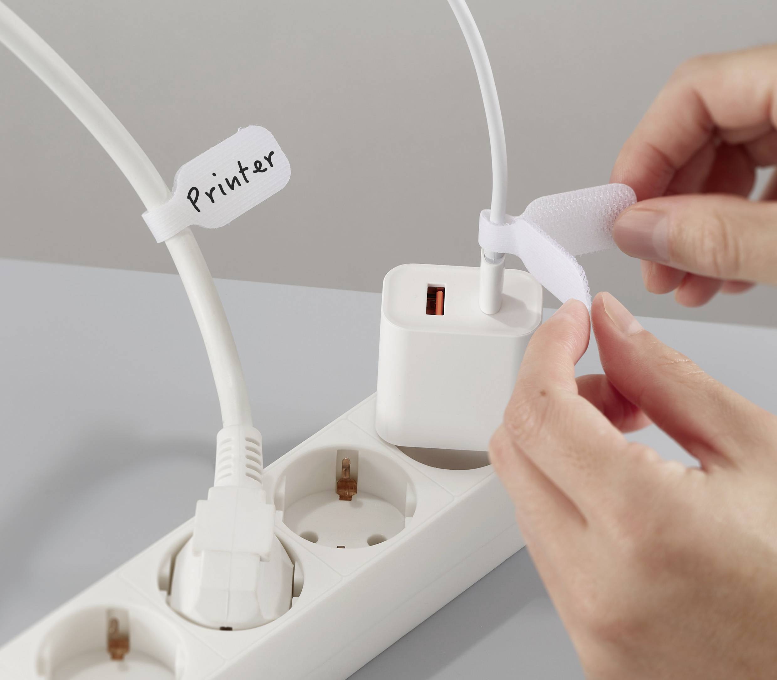 A person is organizing electrical cords on a power strip with a cable marked 'Printer', using a Velcro strap to secure the cables.