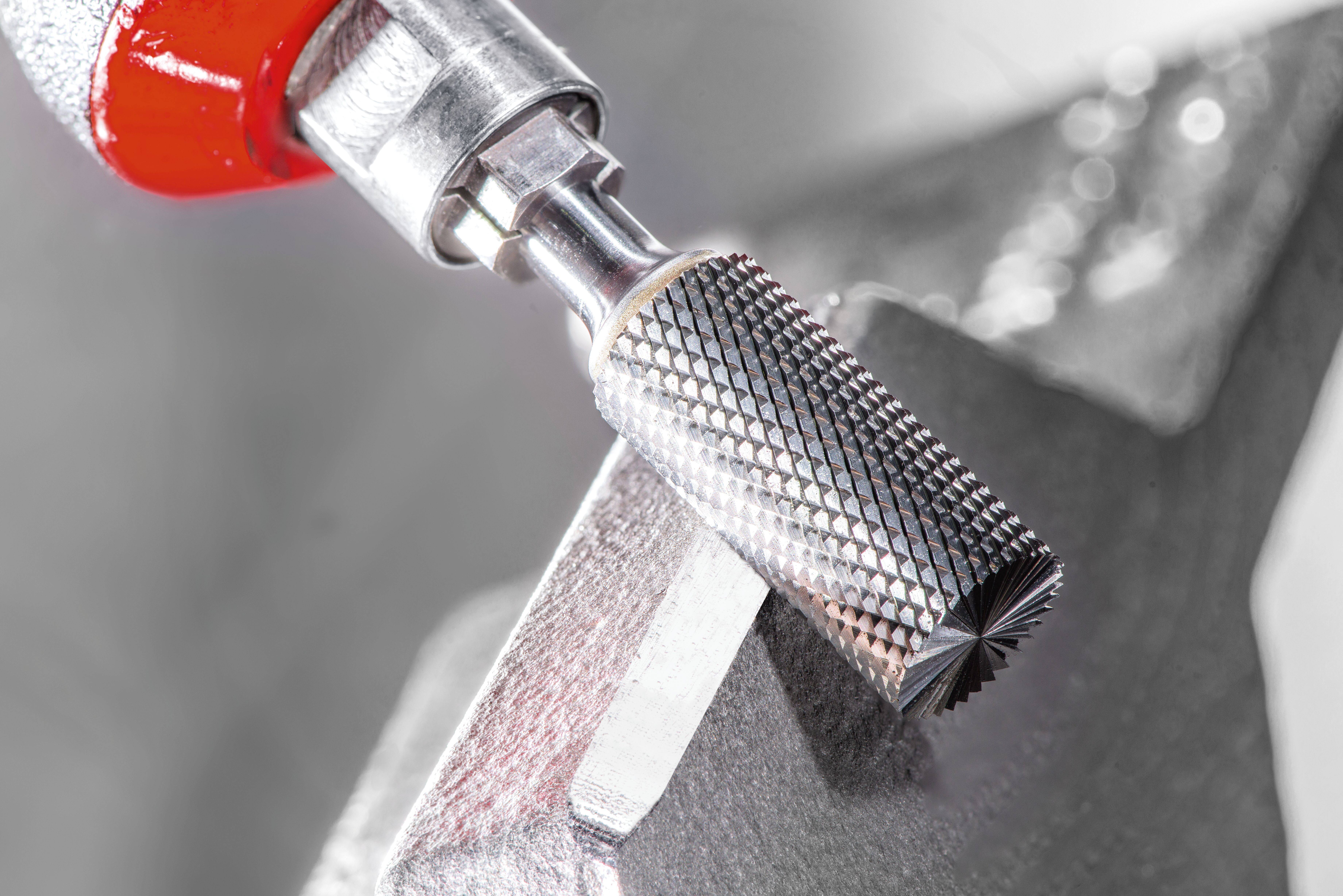 A metal tool with a textured cylindrical tip used for shaping or grinding; it is shown attached to a red holder against a blurred background.