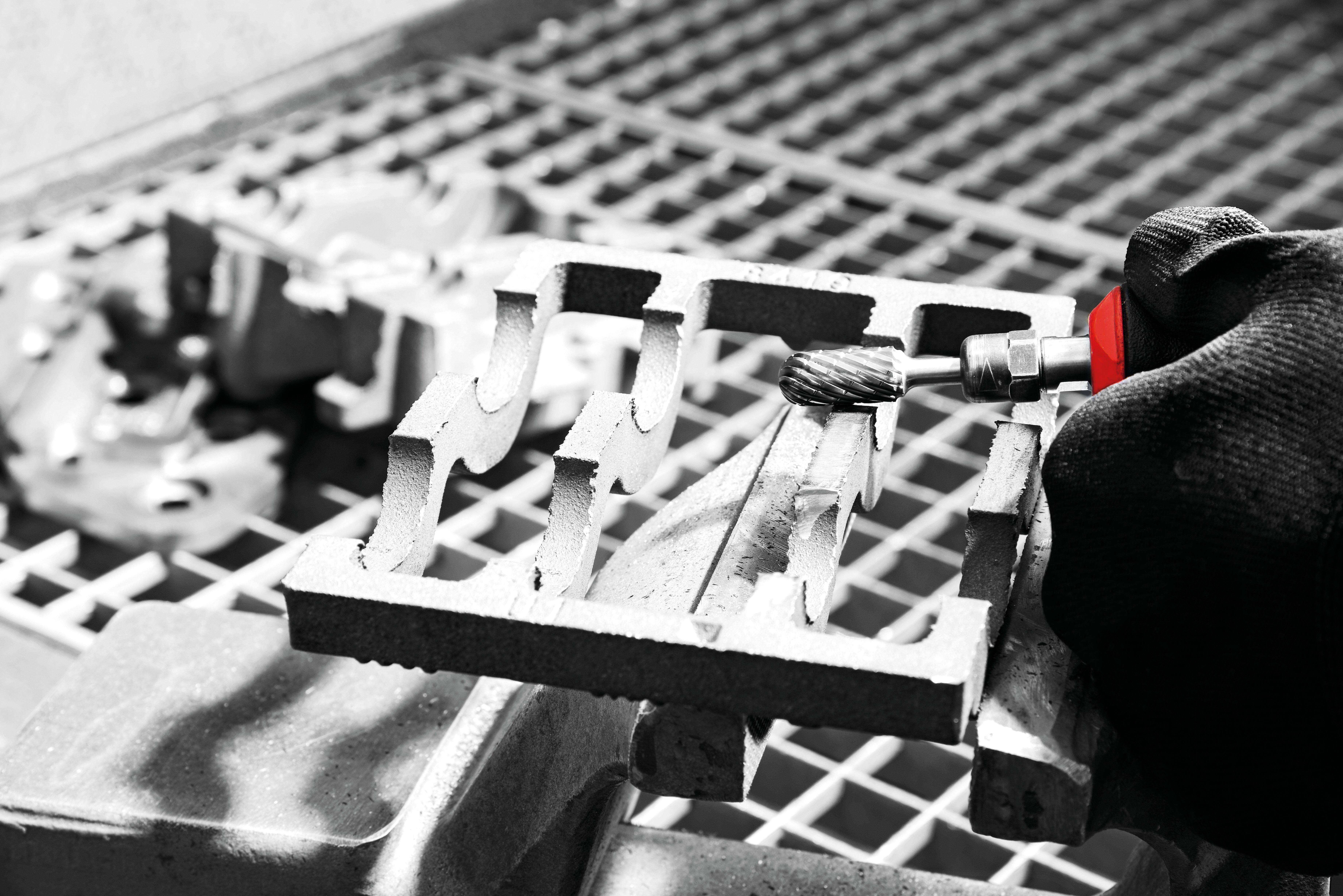 A gloved hand holds a metal stencil against a grated surface while using a rotary tool to smooth the edges.