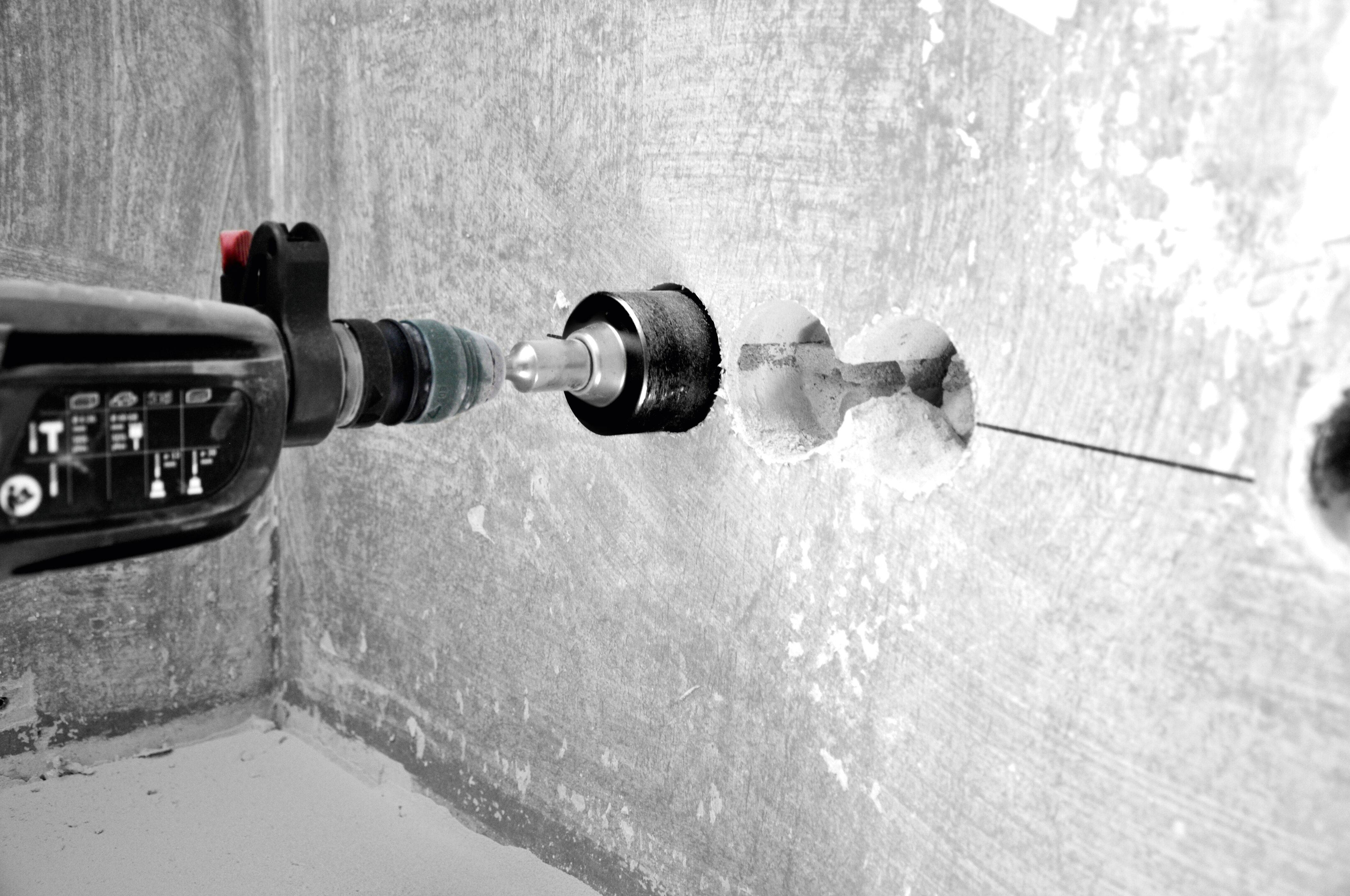 A power drill with a hole saw attachment is creating a circular hole in a concrete wall, showing ongoing construction work.
