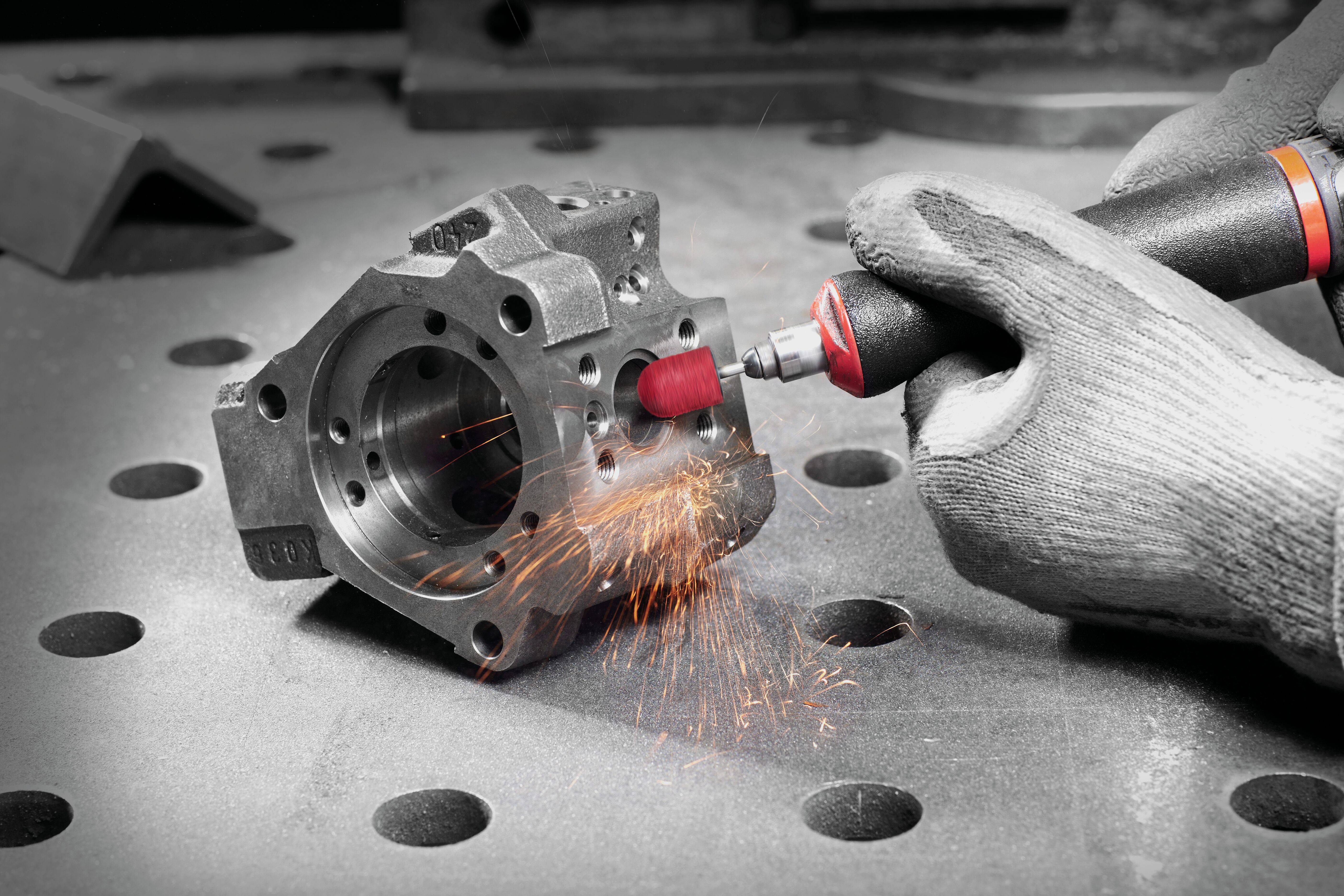 A person wearing gloves uses a rotary tool to grind a metal engine component, with sparks flying from the tool's contact point.