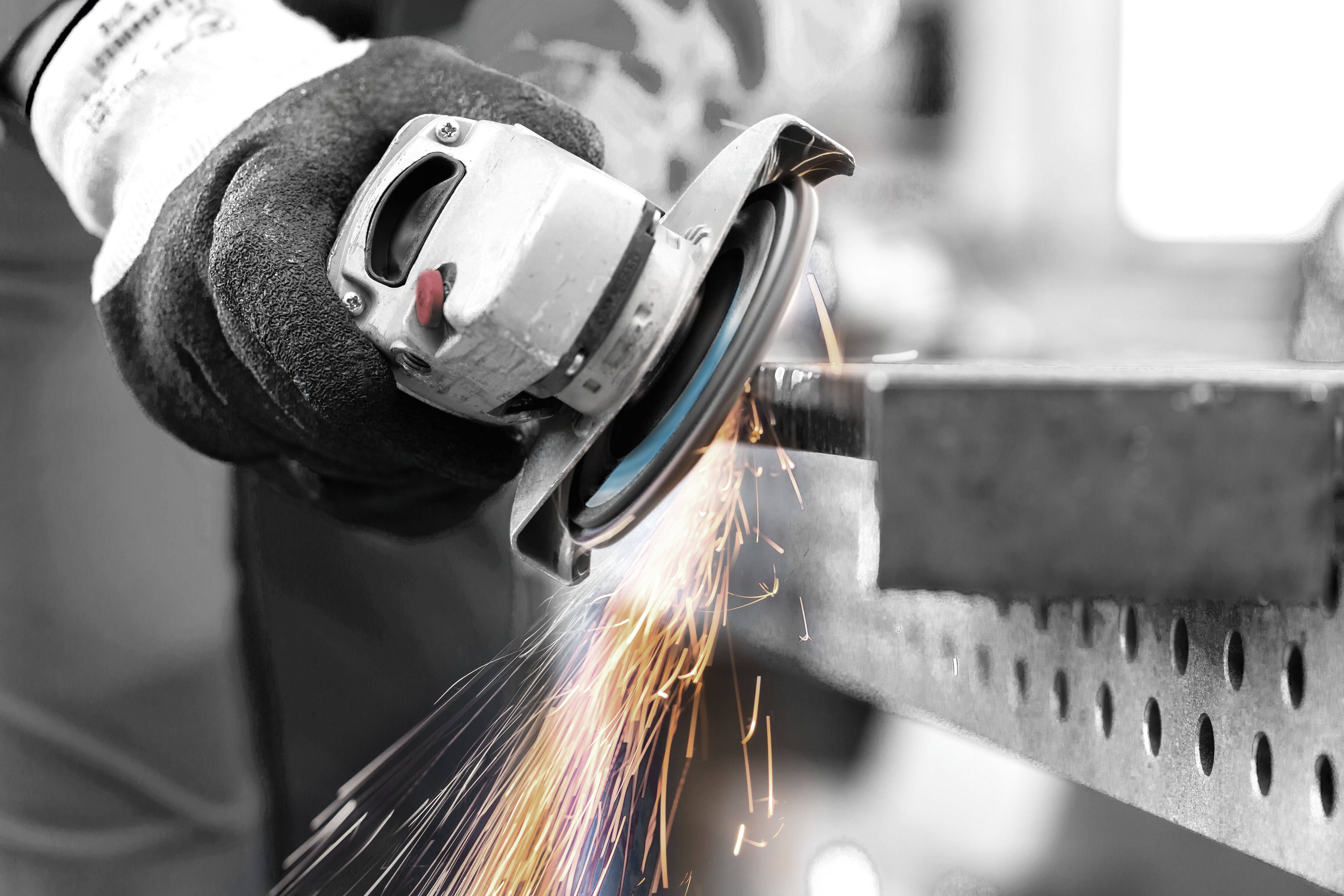 A person in gloves operates an angle grinder, causing bright sparks to fly as it cuts through a metal sheet in an industrial setting.