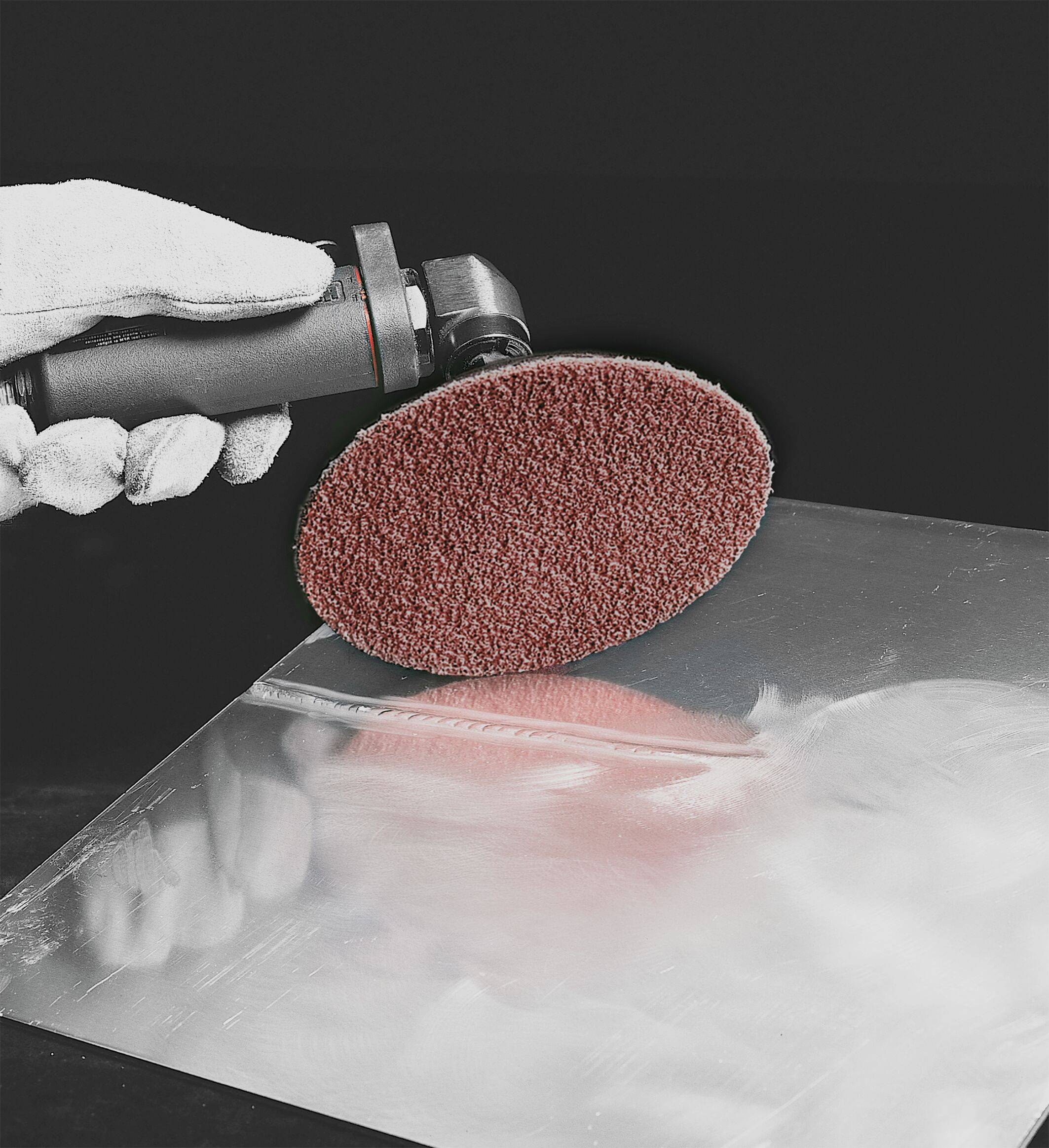 A hand in a white glove holds a power tool with a circular sanding disc, smoothing the surface of a metallic sheet.