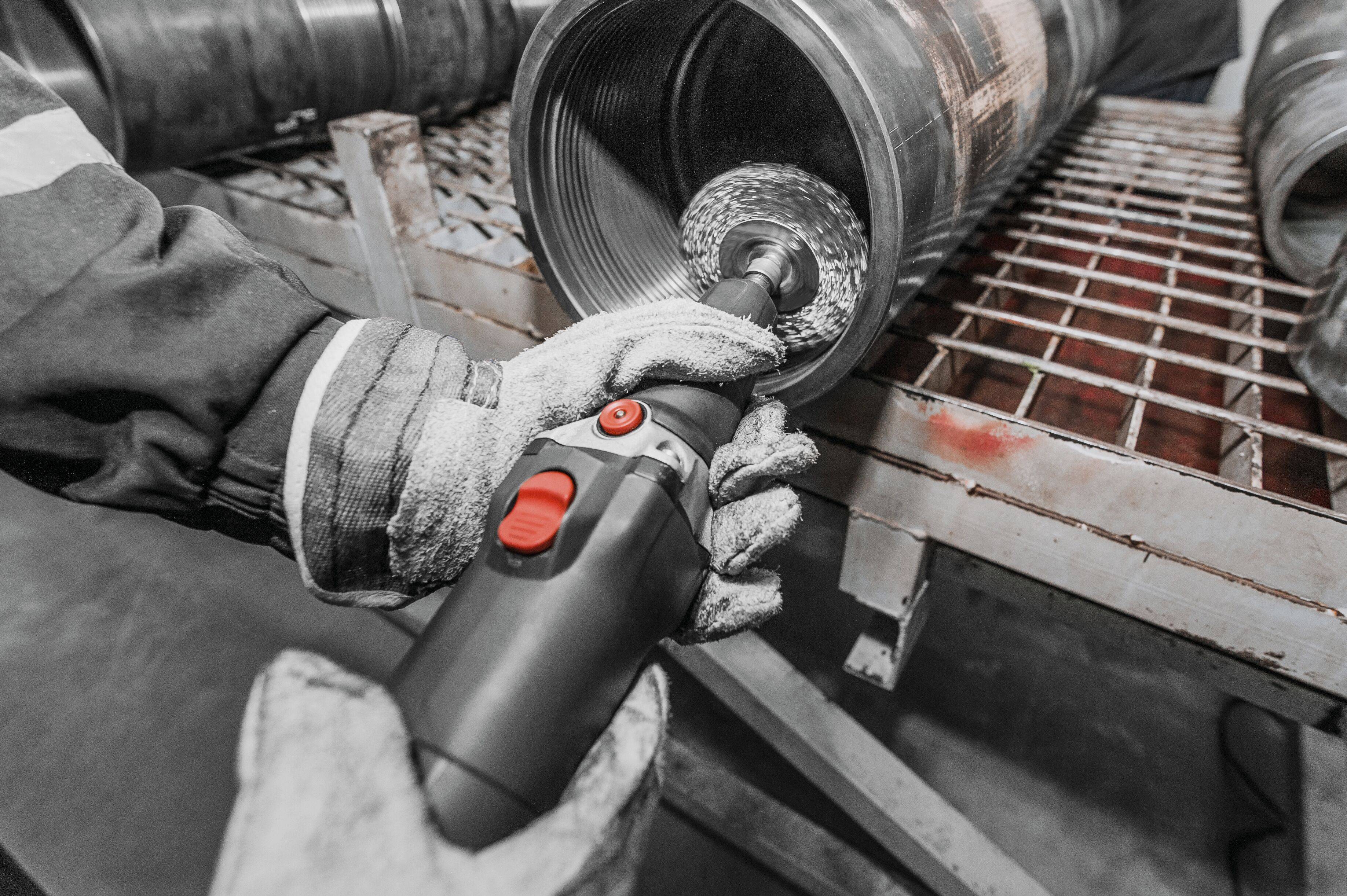 A person wearing protective gloves uses a power tool to polish or clean the inside of a large metal pipe on a workbench.