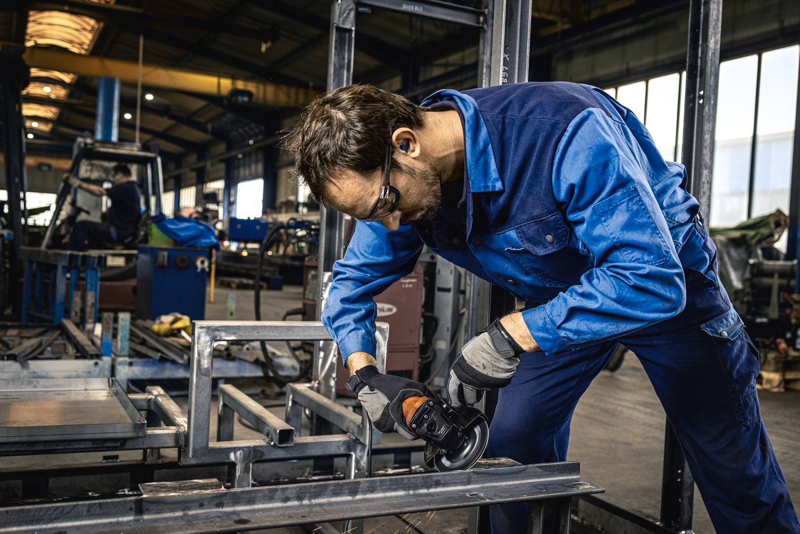 A worker in a blue jumpsuit and safety gear grinds metal in a factory, surrounded by equipment and machinery, focusing on precision.