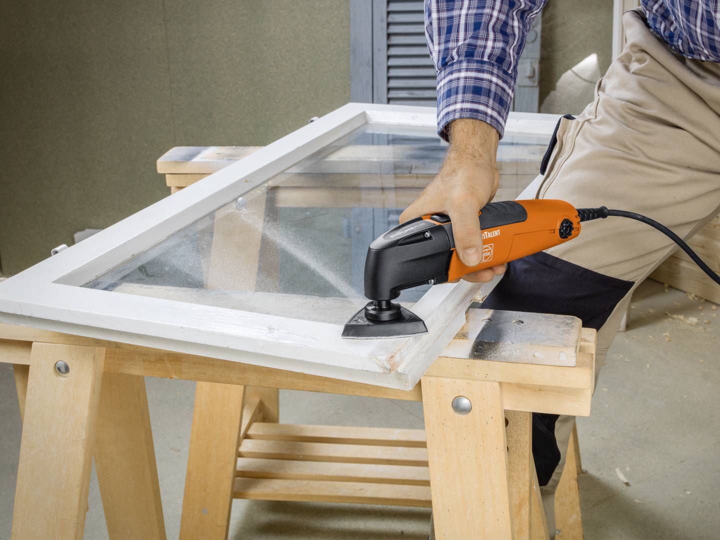 A person uses an orange oscillating tool to sand a wooden window frame resting on a workbench, highlighting a home improvement task.