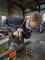 A person wearing safety glasses and a work apron operates a power drill on a metal bar in a workshop. Various drill bits and tools are on the table.
