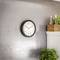 A round wall clock shows 10:09. It hangs on a gray wall above a potted plant, with decorative bottles in the background.