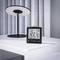 A digital clock displaying '6:30' with temperature readings stands on a white table beside a modern, circular lamp.