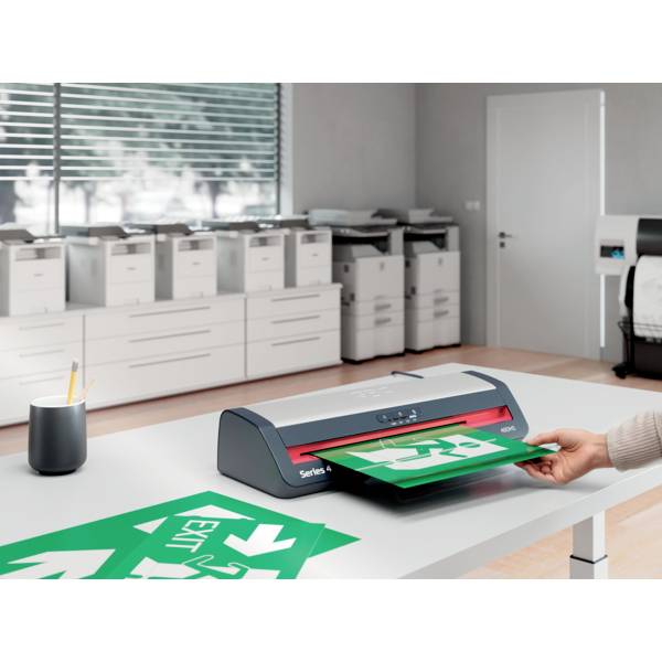 A person uses a laminating machine to laminate green 'EXIT' signs in an office with multiple printers in the background.