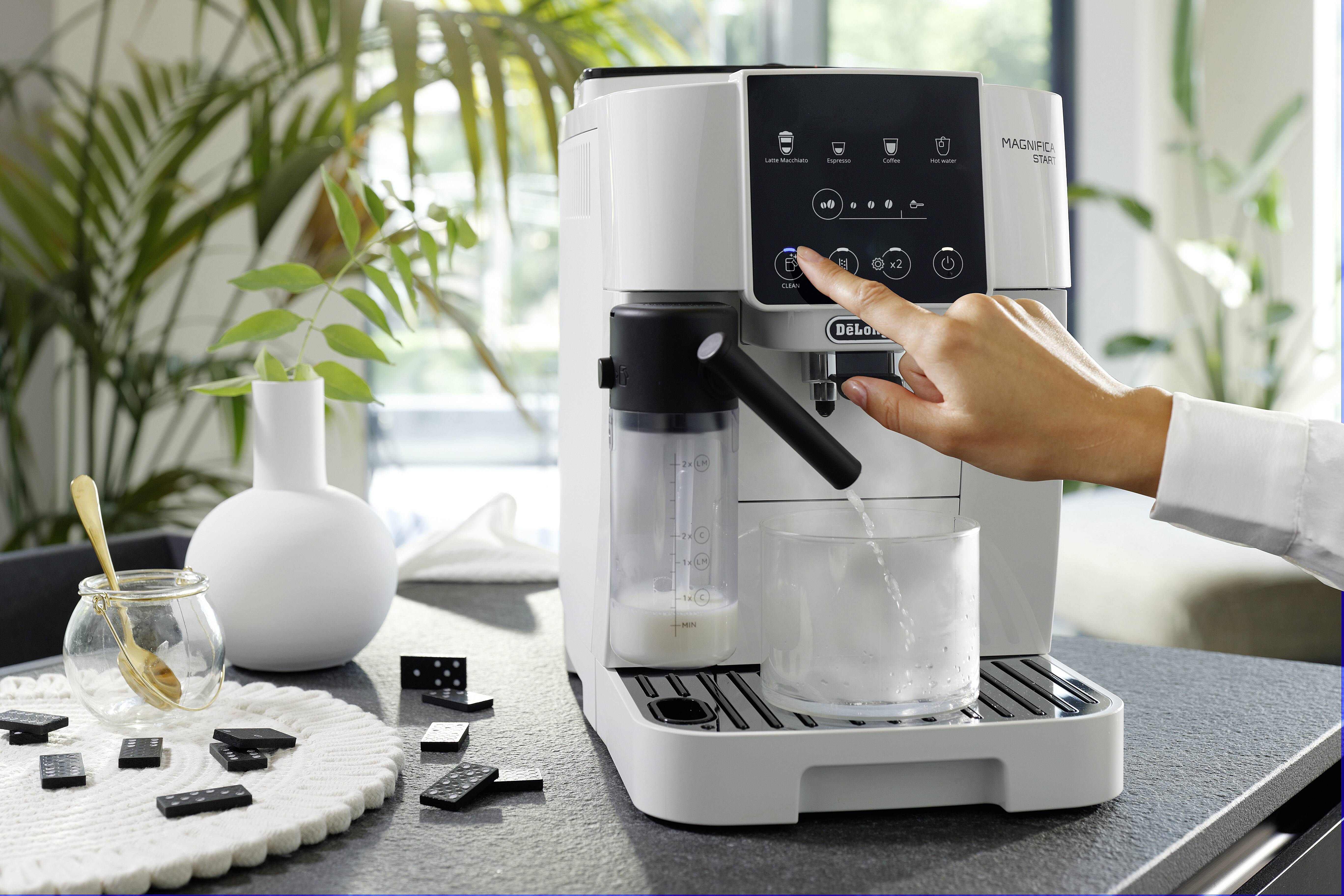 A person's hand uses a modern espresso machine to make coffee. A milk frother and a tray with dominoes are visible on the counter.
