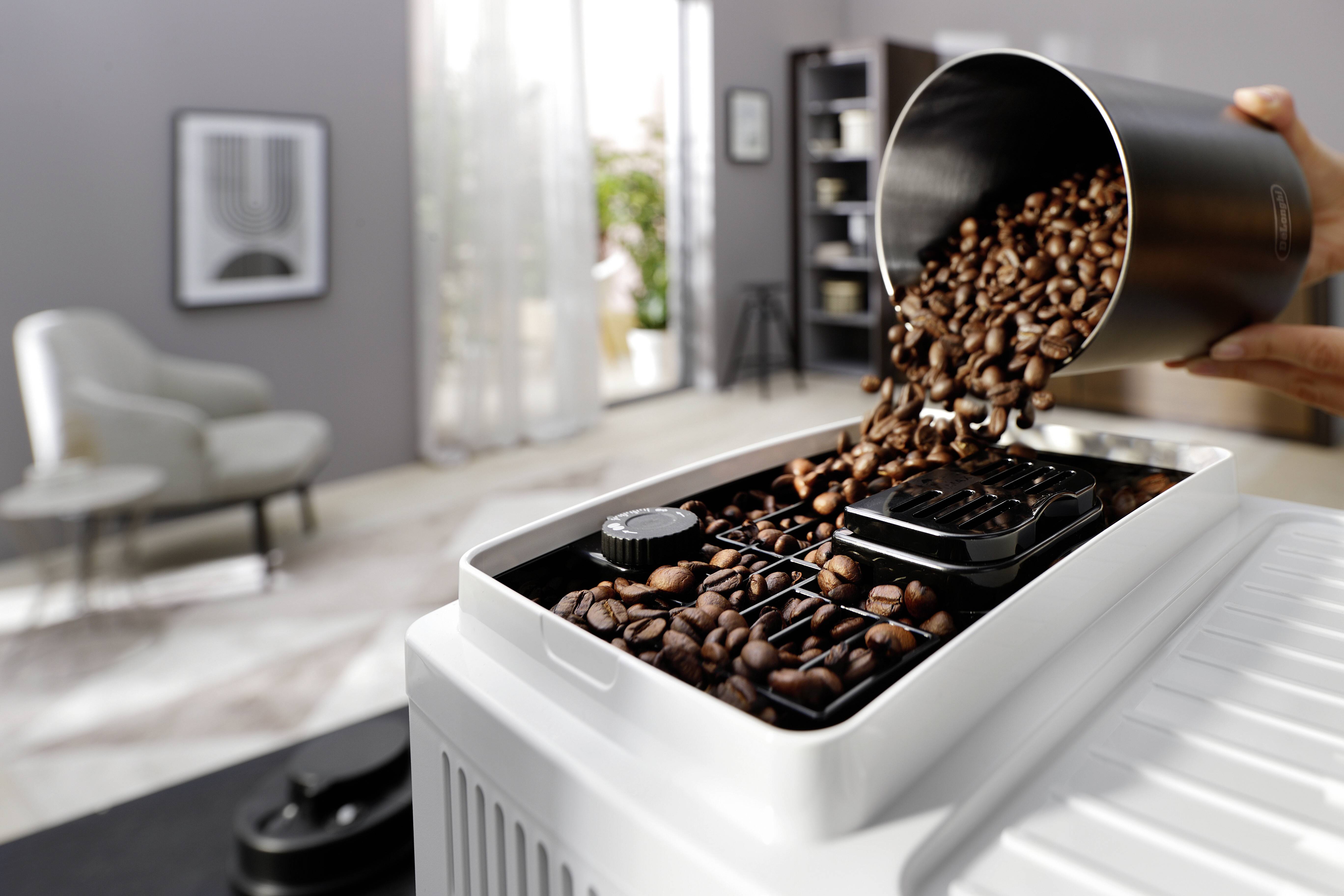 Pouring coffee beans into a white coffee grinder, with a blurred background showing a modern, gray-toned living room.