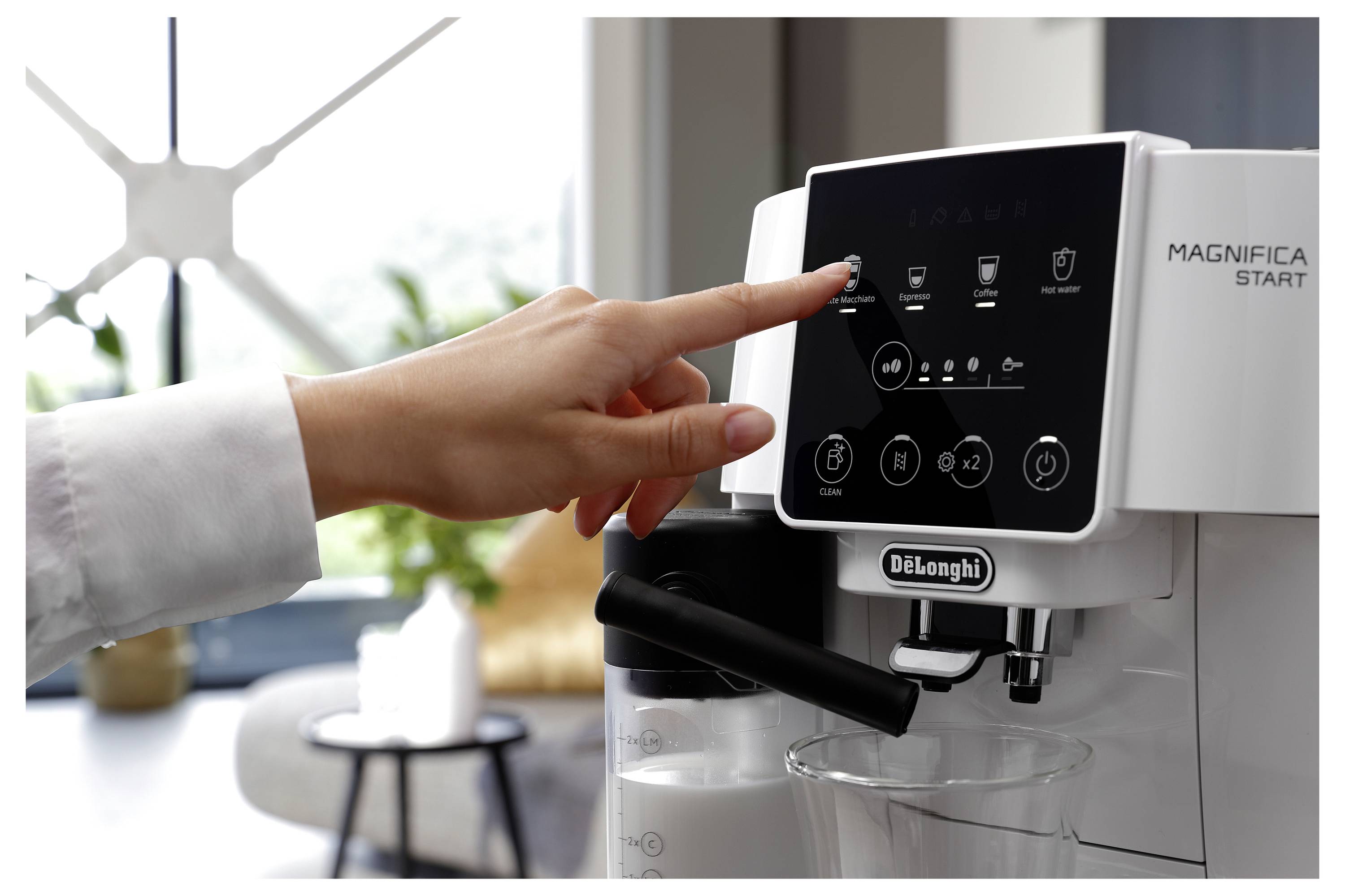 A hand selects a coffee option on a DeLonghi Magnifica Start espresso machine touchscreen, with a modern living room in the background.