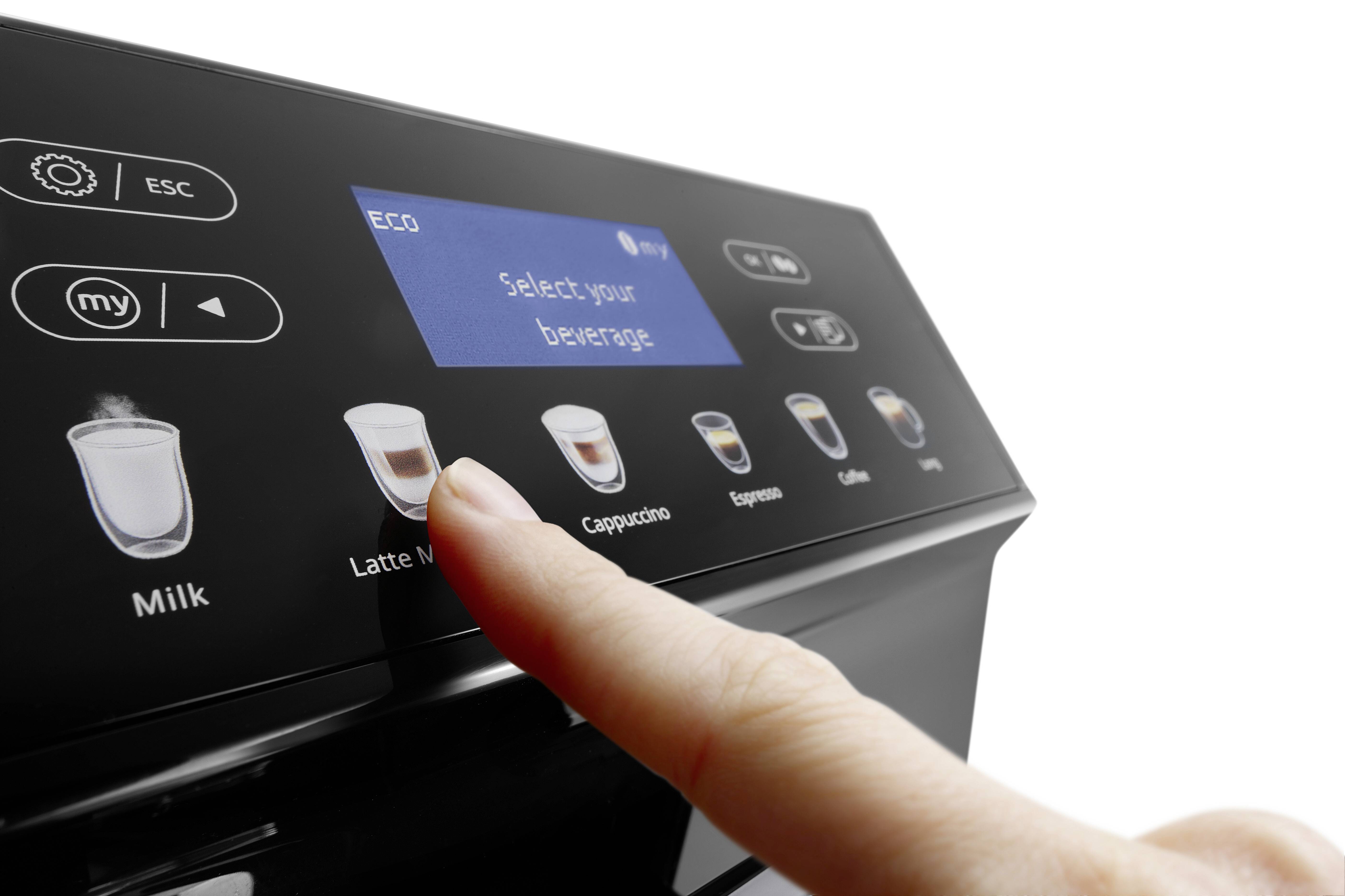 A finger presses the 'Latte Macchiato' option on a coffee machine touch panel, with various coffee types displayed on the screen.