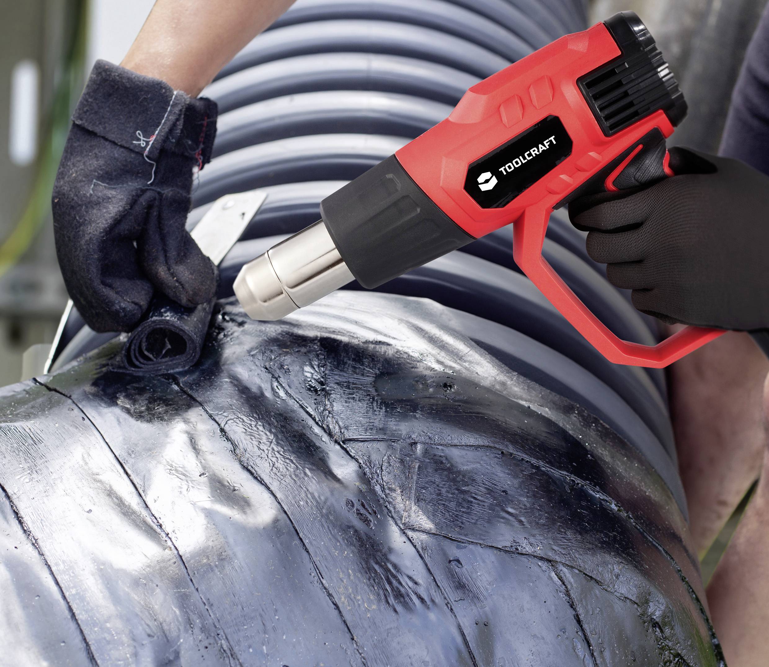 'Person using a red heat gun to fuse or seal layers of a large, metallic or plastic pipe; wearing gloves for protection.'