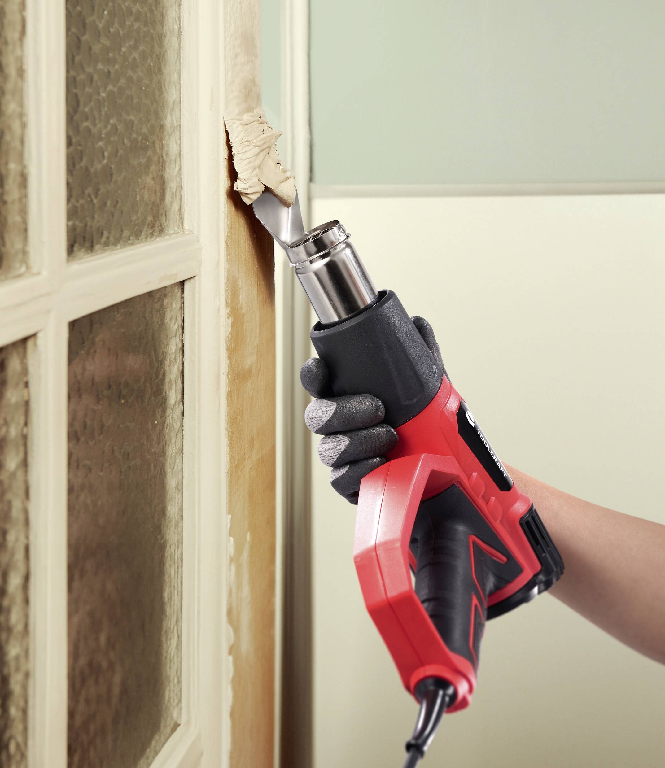 A person uses a red and black heat gun to remove paint from a wooden door frame, highlighting DIY renovation work.
