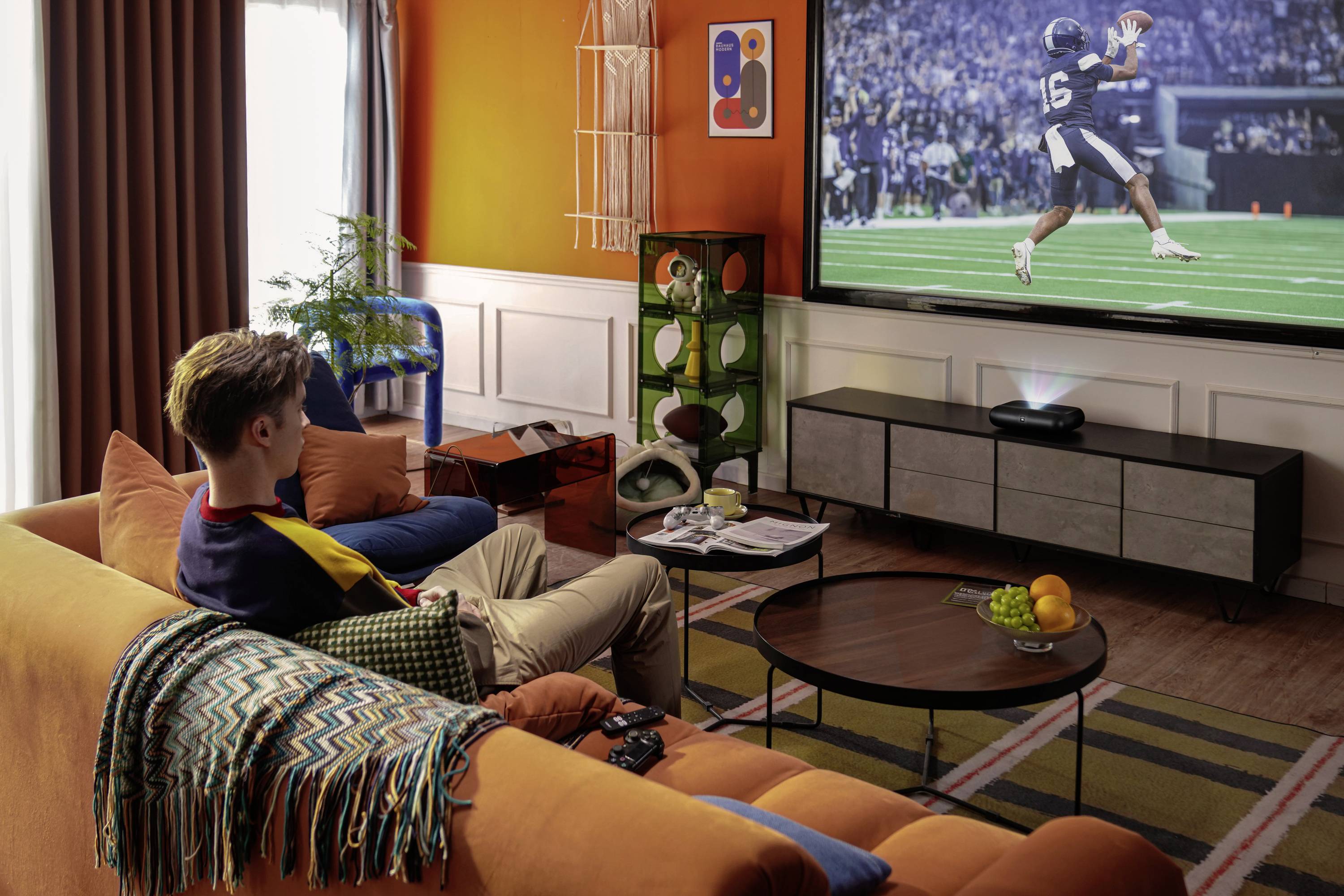 A person sitting on an orange sofa watches a football game on a large TV screen in a cozy living room with vibrant decor.