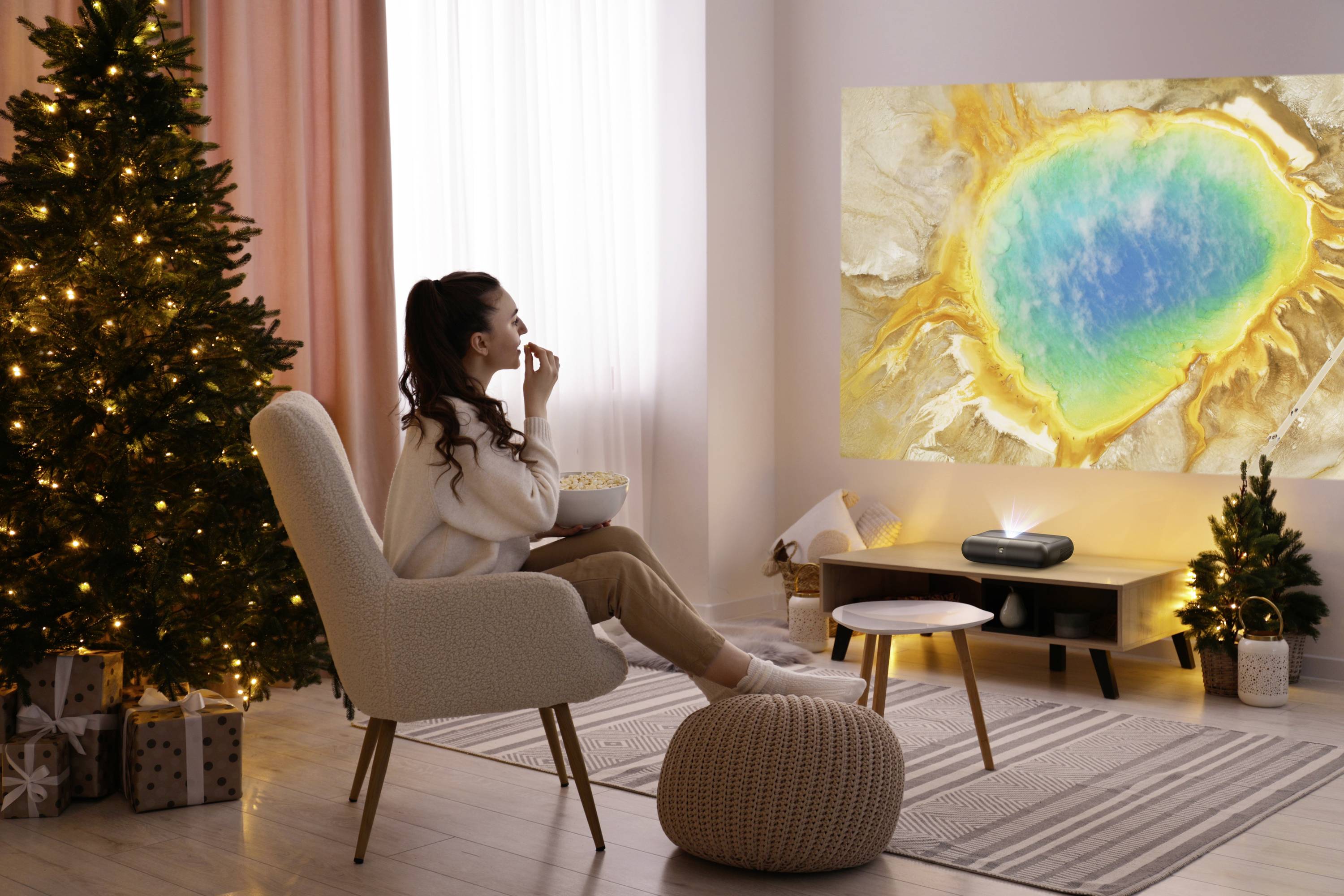 A woman sits in a cozy room, watching a vibrant natural scene projected on the wall, with a decorated Christmas tree nearby.