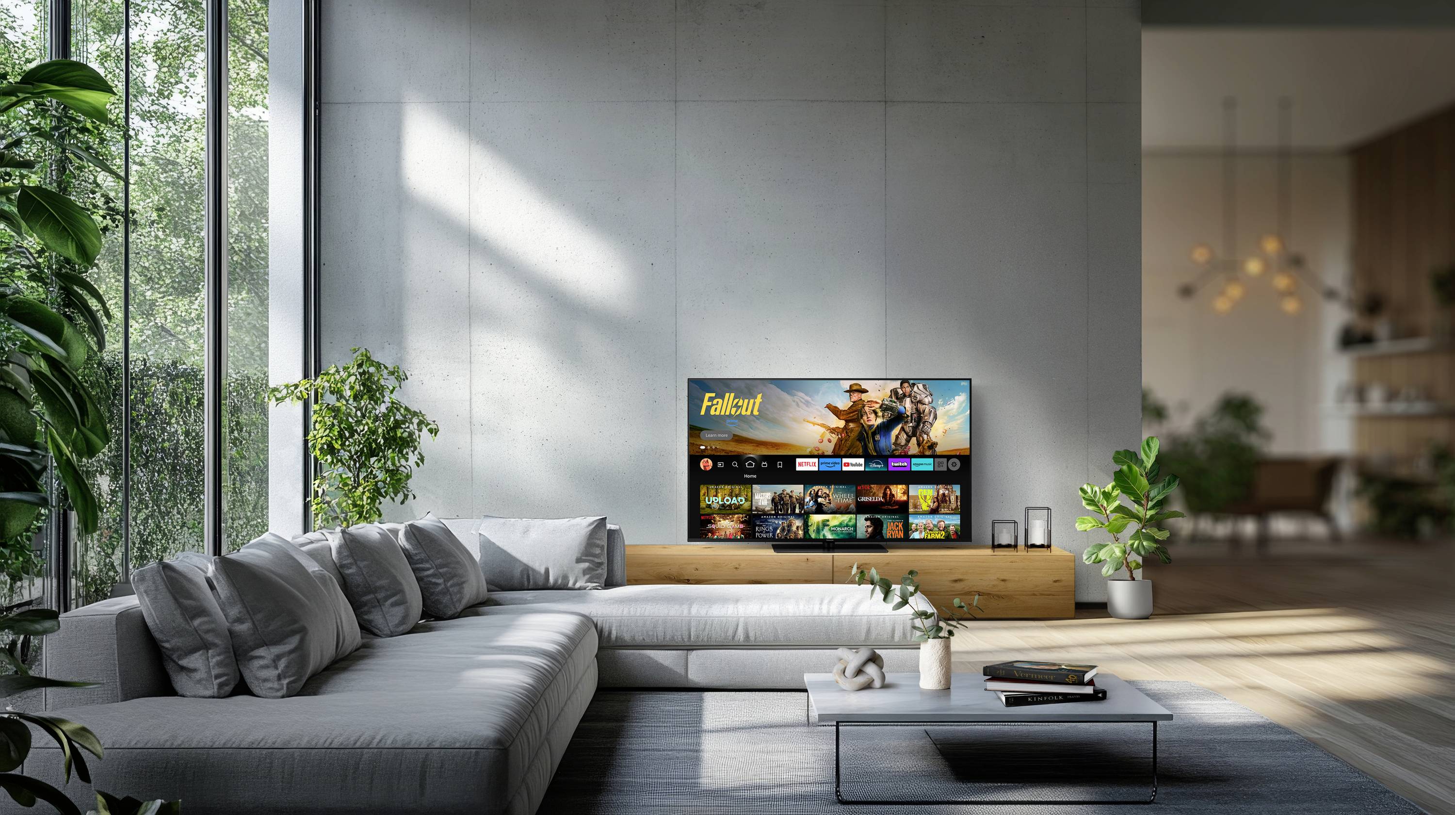 A modern living room with a large window showing greenery outside. A wide gray sofa faces a flat-screen TV displaying streaming content.