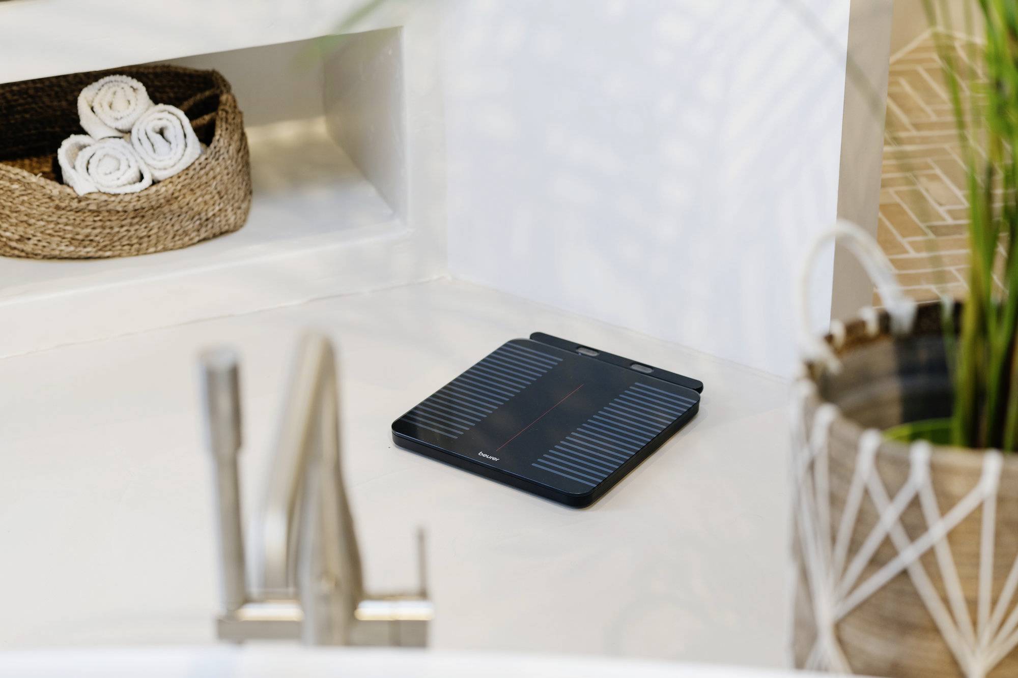 A bathroom scale on a white floor near a plant and a basket of rolled towels in a modern bathroom setting.
