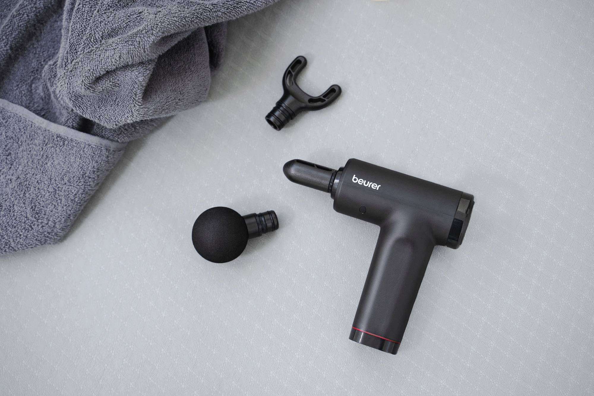 A black massage gun with attachments lies on a light surface next to a gray towel.