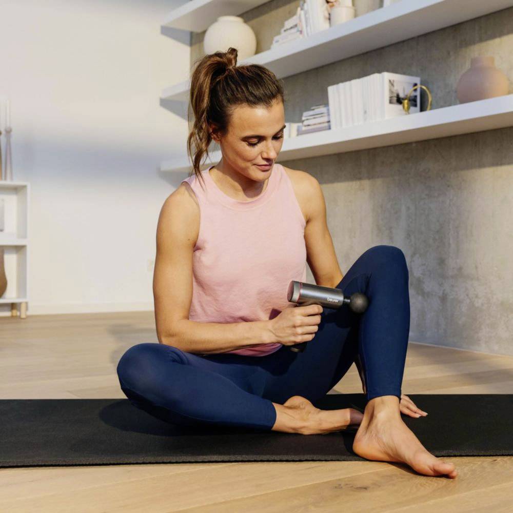 A woman sitting on a yoga mat uses a massage gun on her leg. Shelves with decor items are in the background.