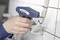 A person uses a blue cable tie gun to secure white cables against a white tiled wall, demonstrating cable management.