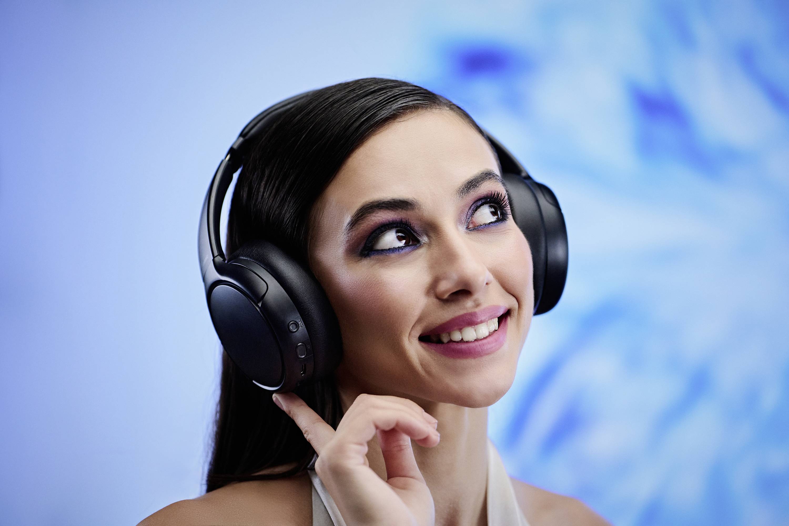 A woman wearing black headphones smiles and looks upward, set against a blurred, abstract blue background.