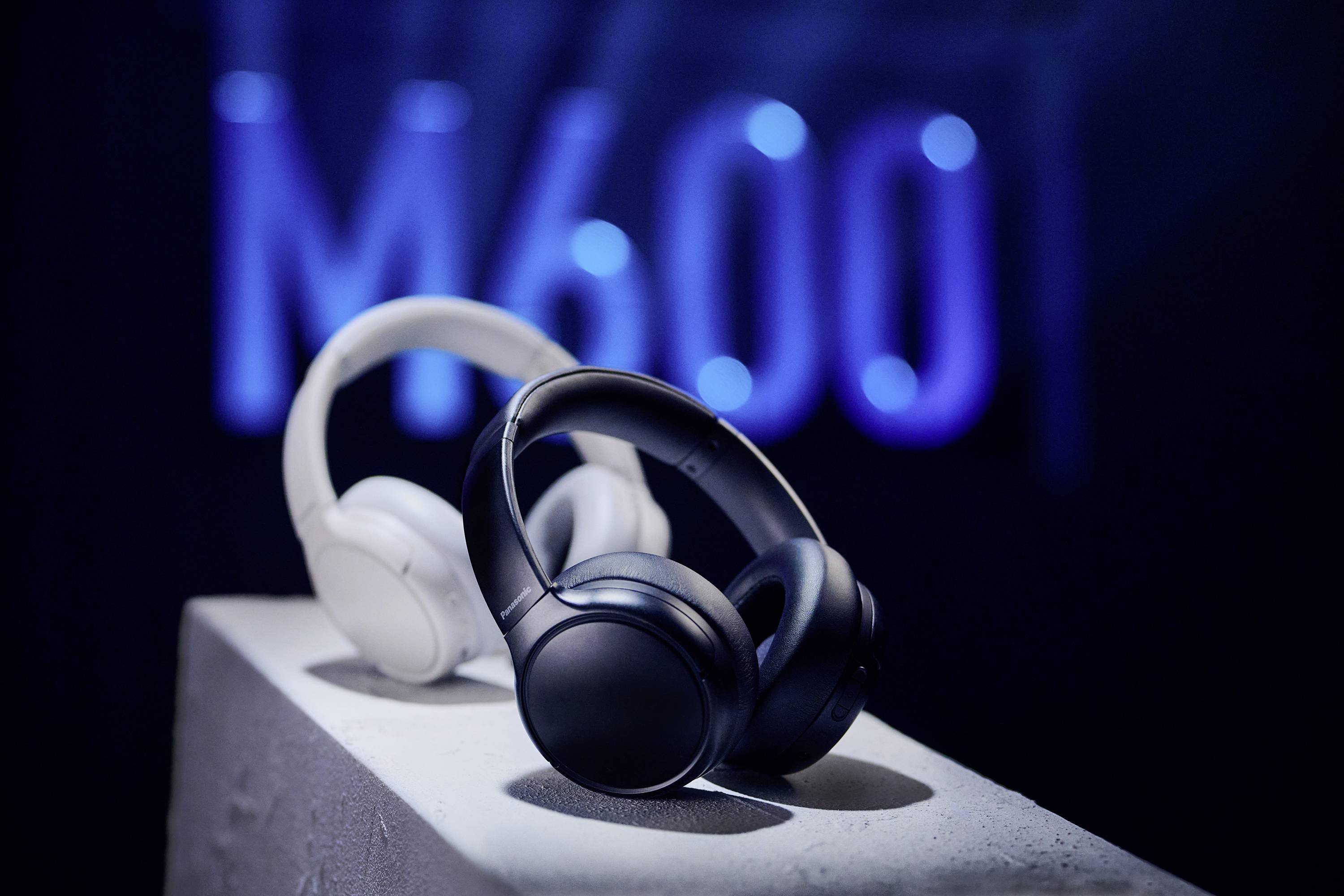 Two pairs of headphones, black and white, are displayed on a pedestal with a blurred 'M600' in blue light in the background.