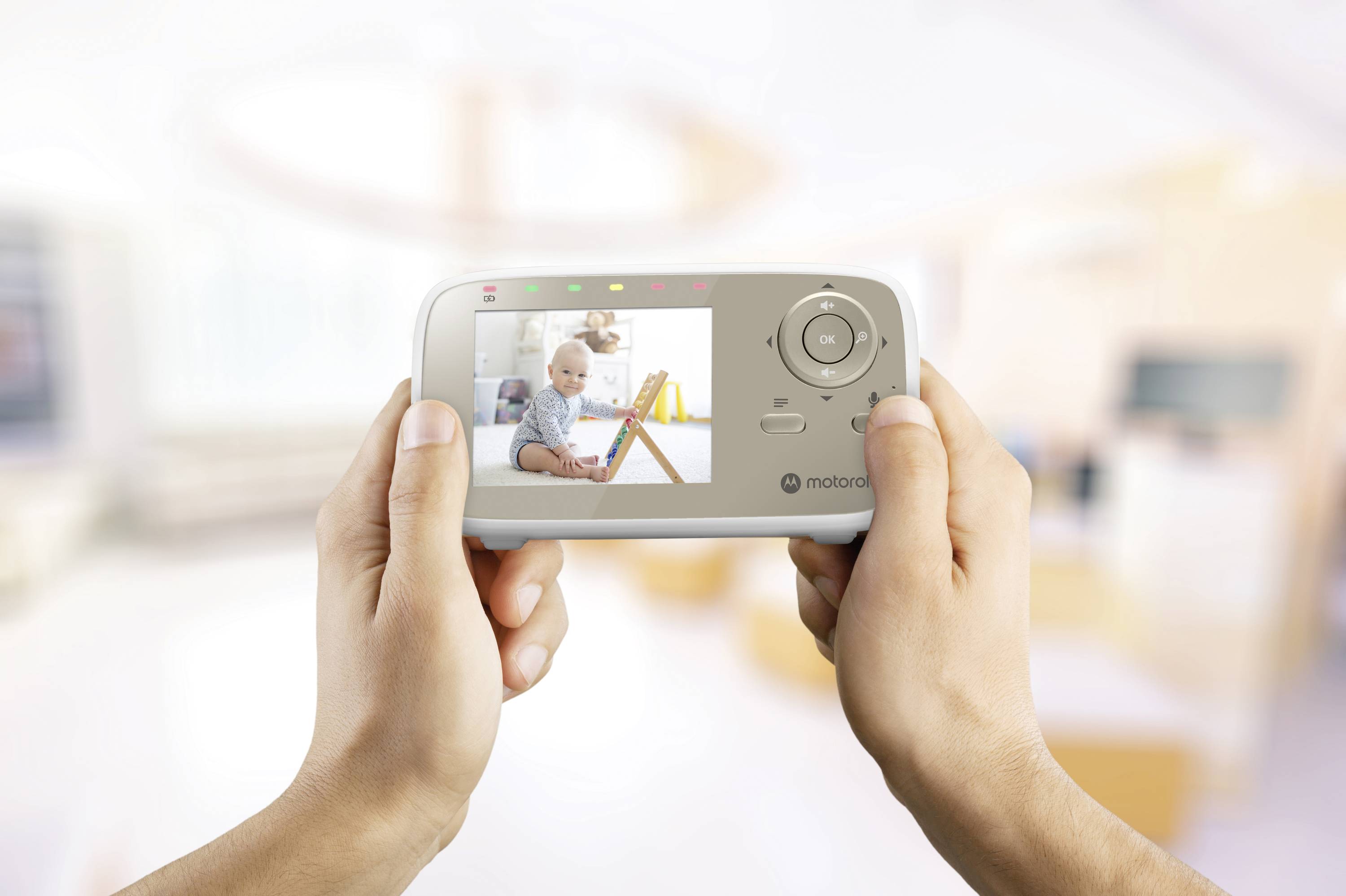 Hands hold a baby monitor showing a sitting baby in a brightly lit room, indicating a safe and monitored environment.