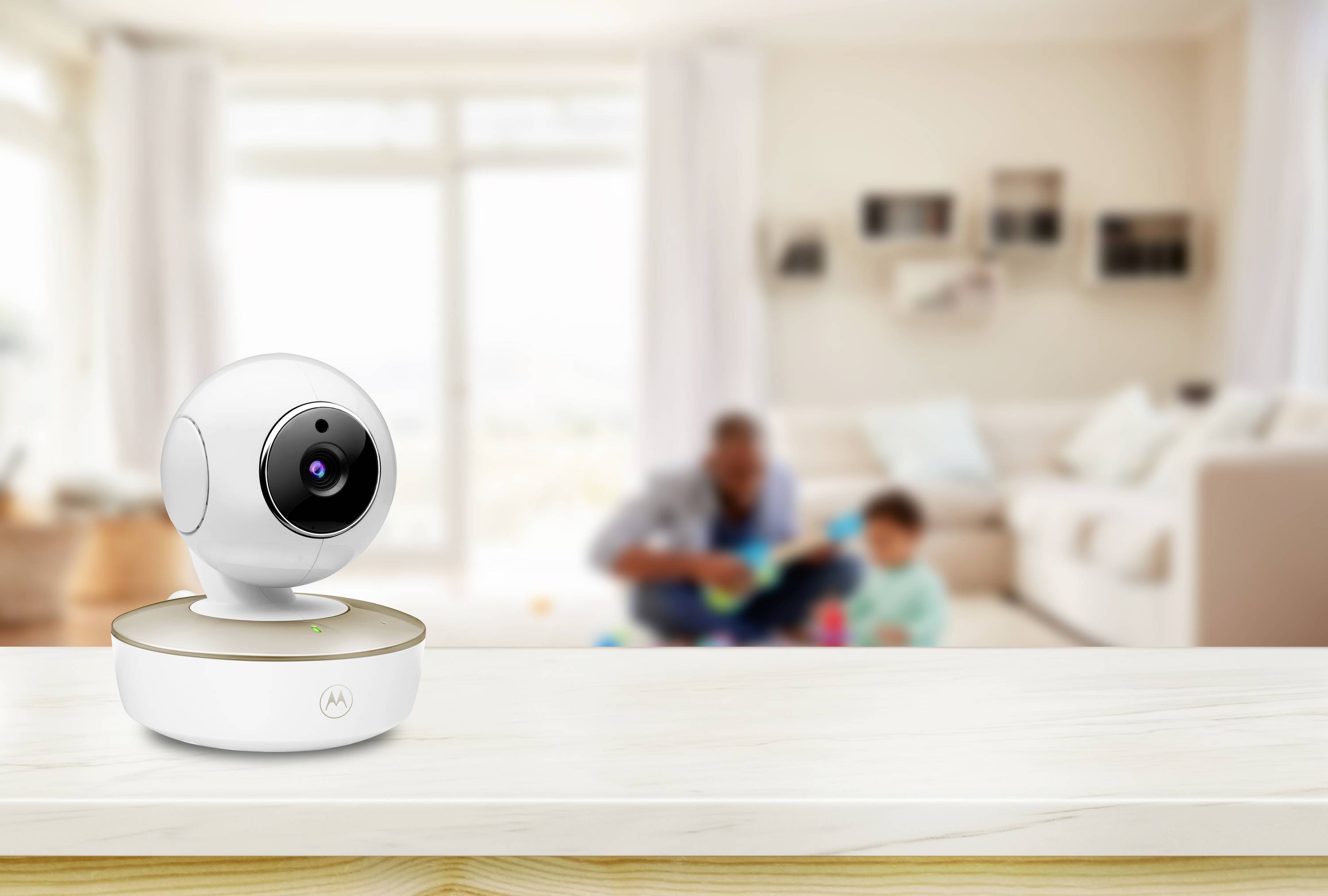 A modern indoor security camera sits on a wooden surface in a bright living room. In the blurred background, an adult and child play.